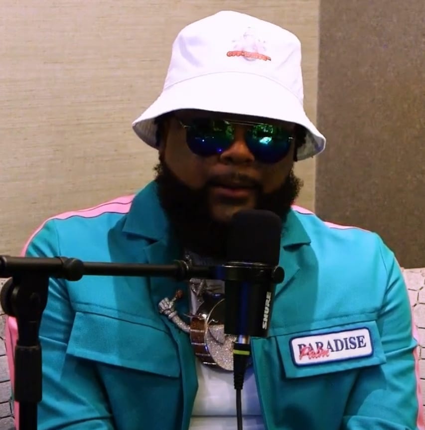 03 Greedo, wearing a white bucket hat, reflective sunglasses and a teal jacket with a Paradise patch, speaks into a microphone during an interview.