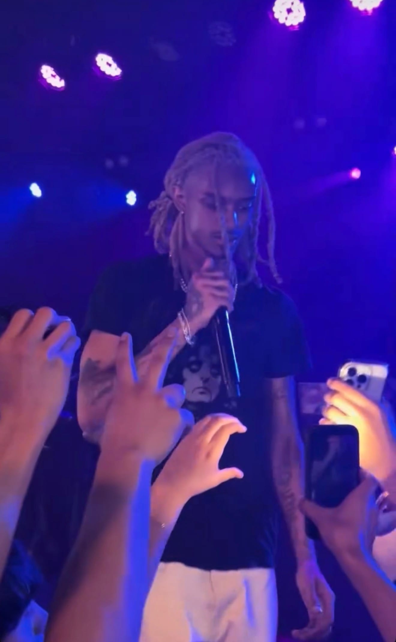 1300SAINT, with bleached dreadlocks and face tattoos, sings into a microphone above a crowd reaching up with phones.