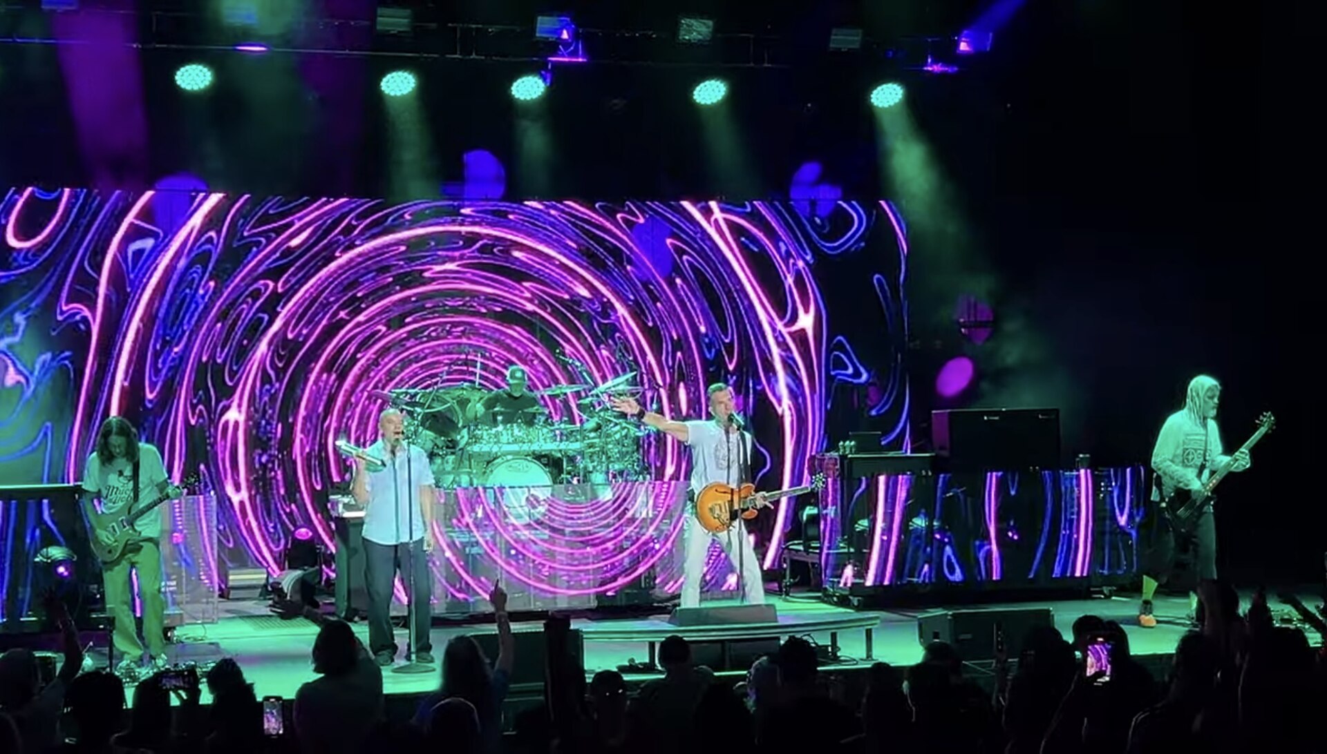 The five members of 311 play across a wide stage in front of a screen of pink and blue spiral graphics, drummer raised on a riser behind them.