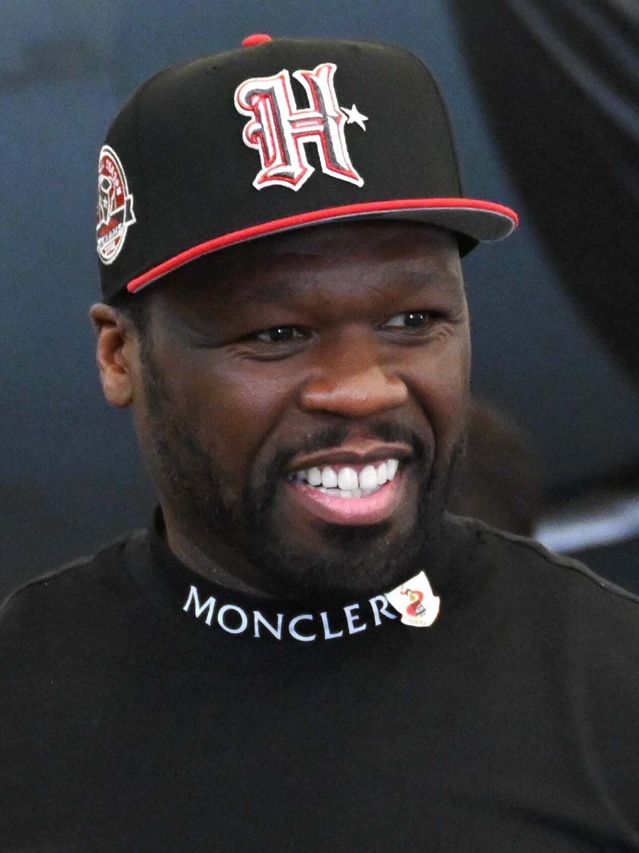A close-up of 50 Cent smiling broadly, wearing a black cap with an 'H' logo and a black Moncler top.
