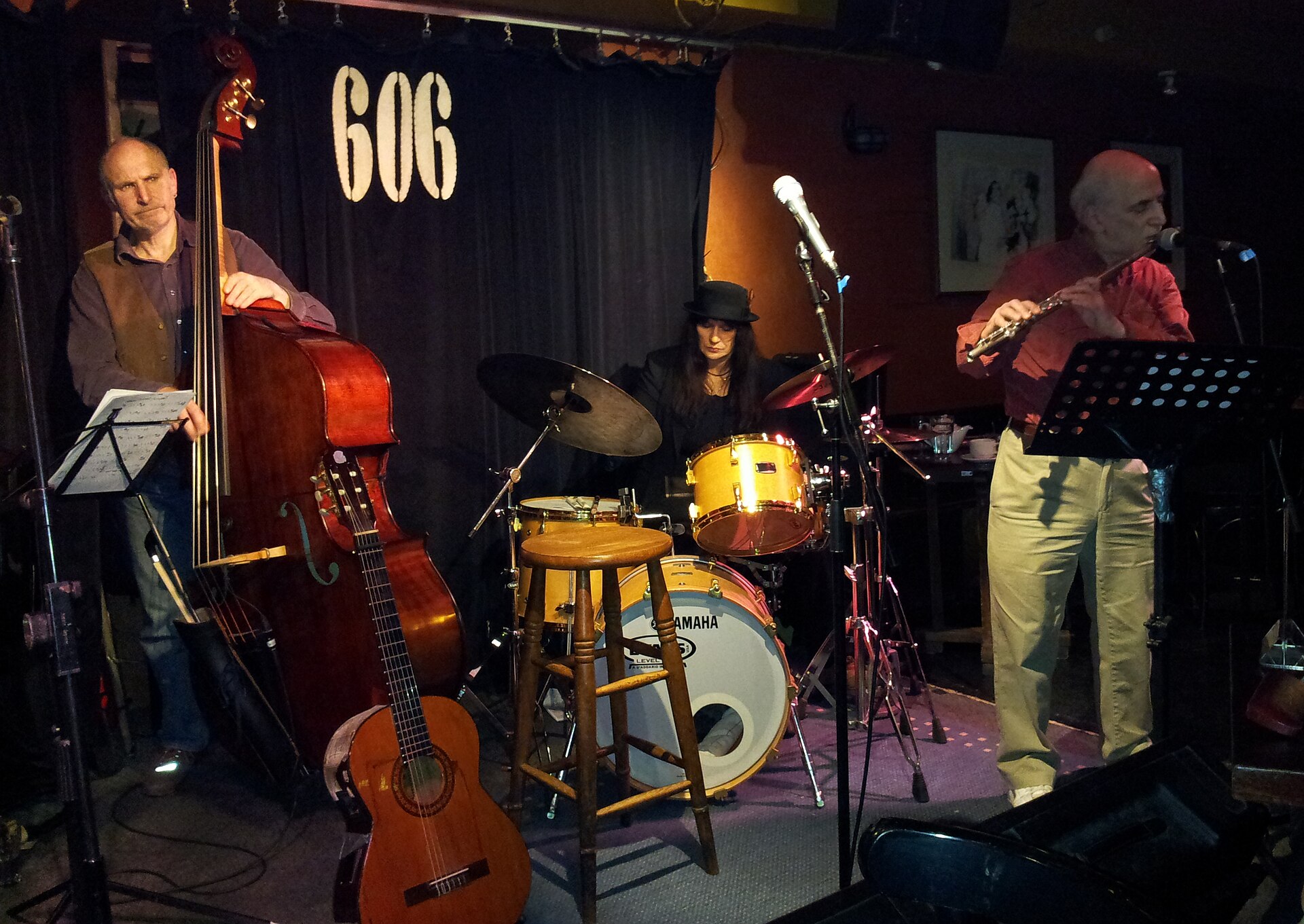 A jazz trio plays in a small basement room under a curtain marked 606 - double bass at left, drums in the middle, flute at right.