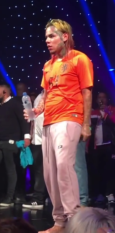 6ix9ine stands on a stage in an orange football shirt and pink track pants, his multicoloured hair tied up on top of his head.
