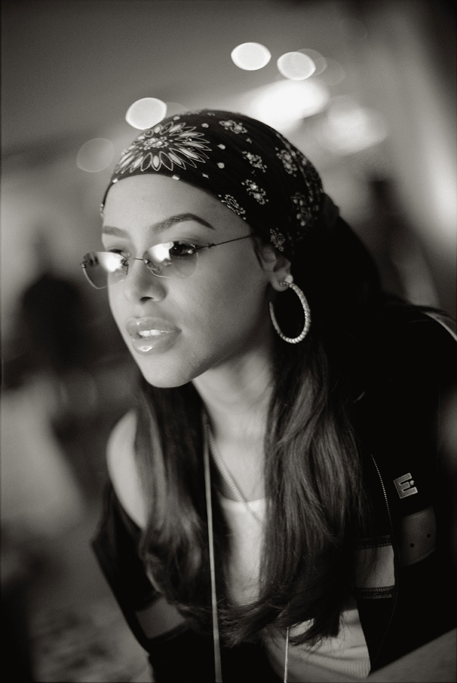 Aaliyah looks toward the camera in a patterned bandana, rimless tinted glasses and a large hoop earring.