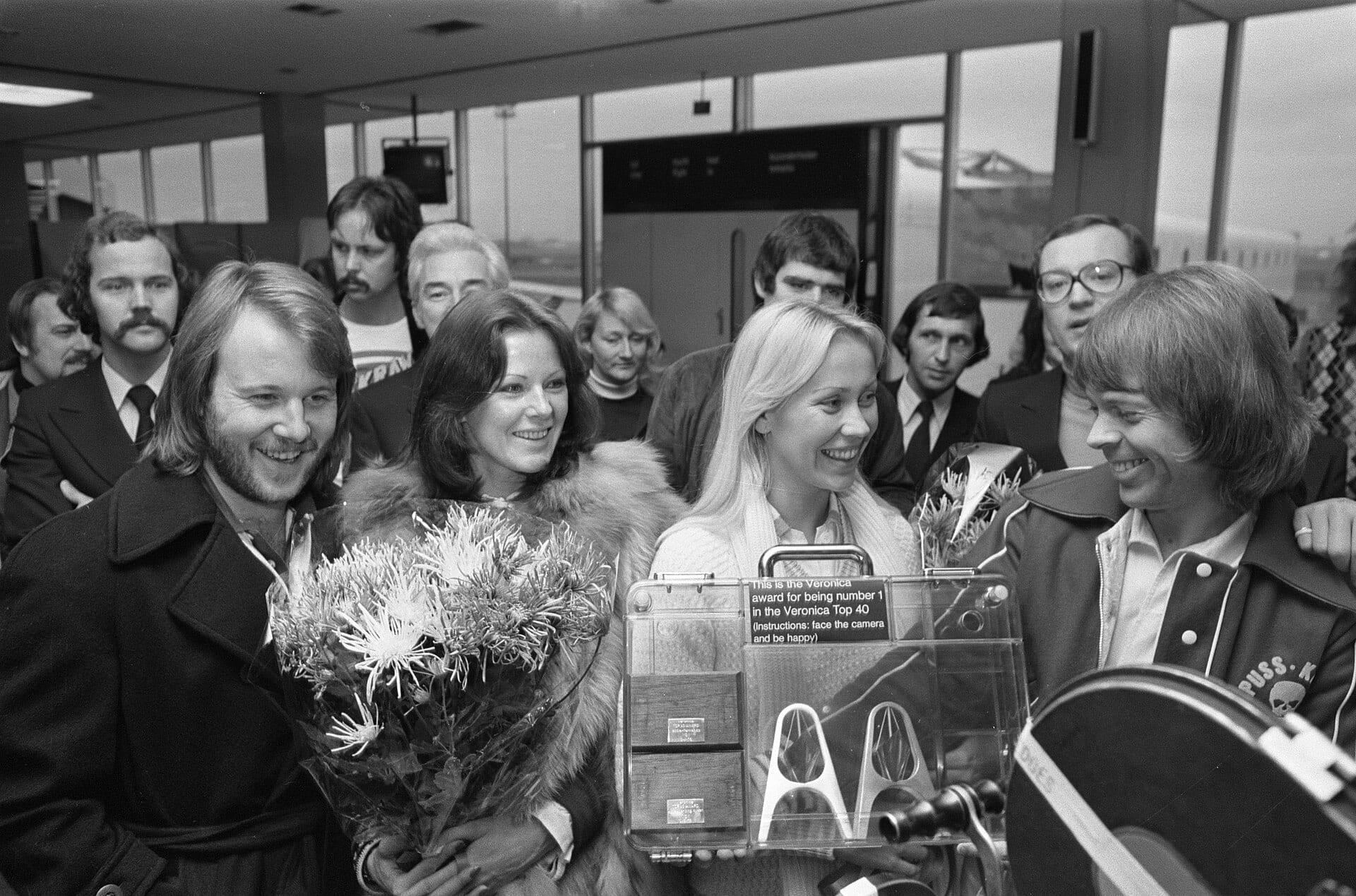 The four members of ABBA smile together at an airport, Benny and Anni-Frid holding a bouquet of flowers while Agnetha holds a display case reading it is the Veronica award for being number 1 in the Veronica Top 40.