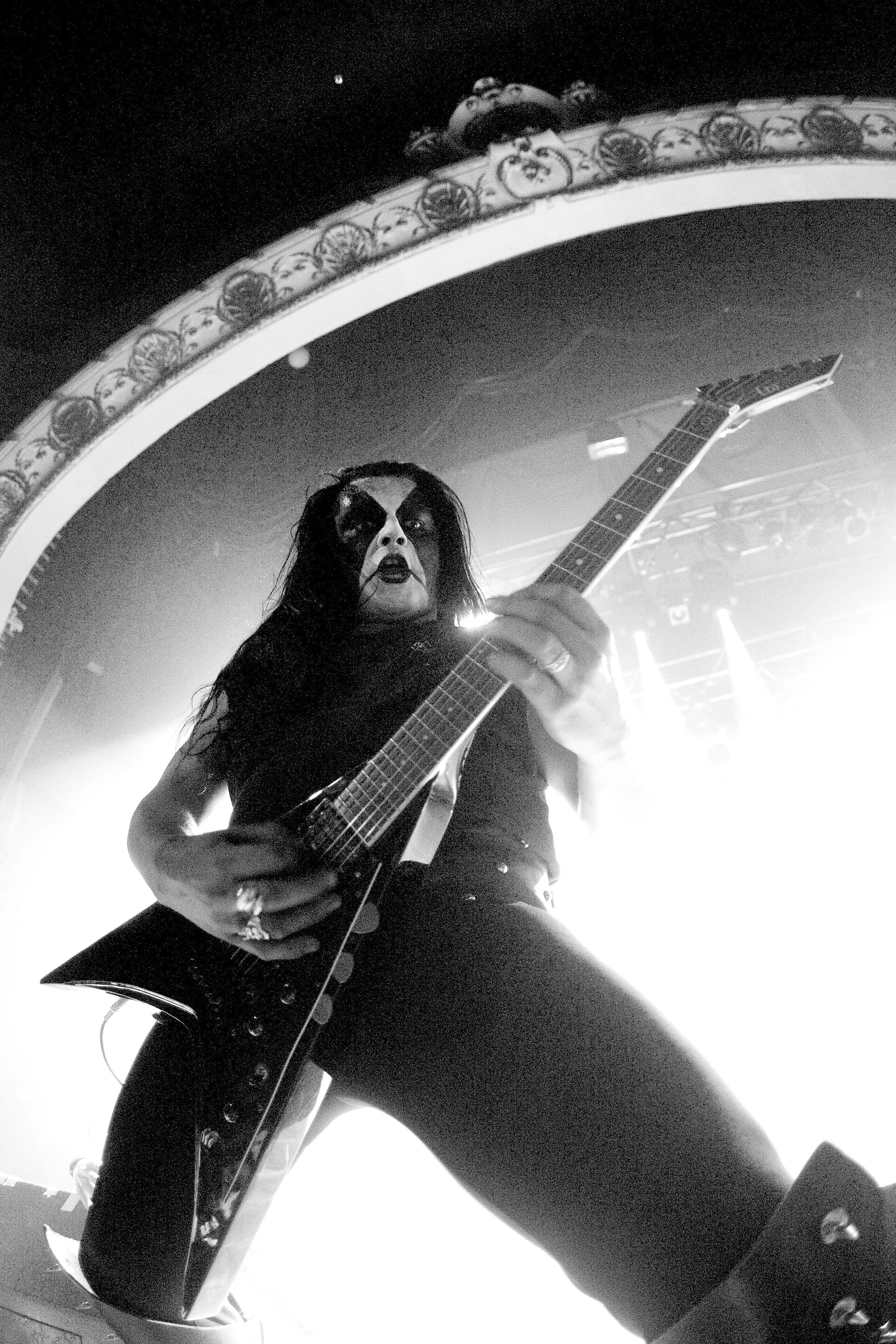 Abbath performs on stage in black and white, wearing black corpse paint and playing a black V-shaped guitar under bright backlighting.