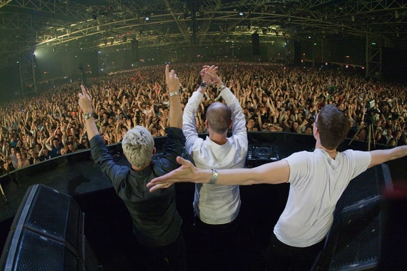 Three members of Above & Beyond stand at a DJ booth with arms raised, backs to the camera, facing a packed arena crowd with hands in the air.