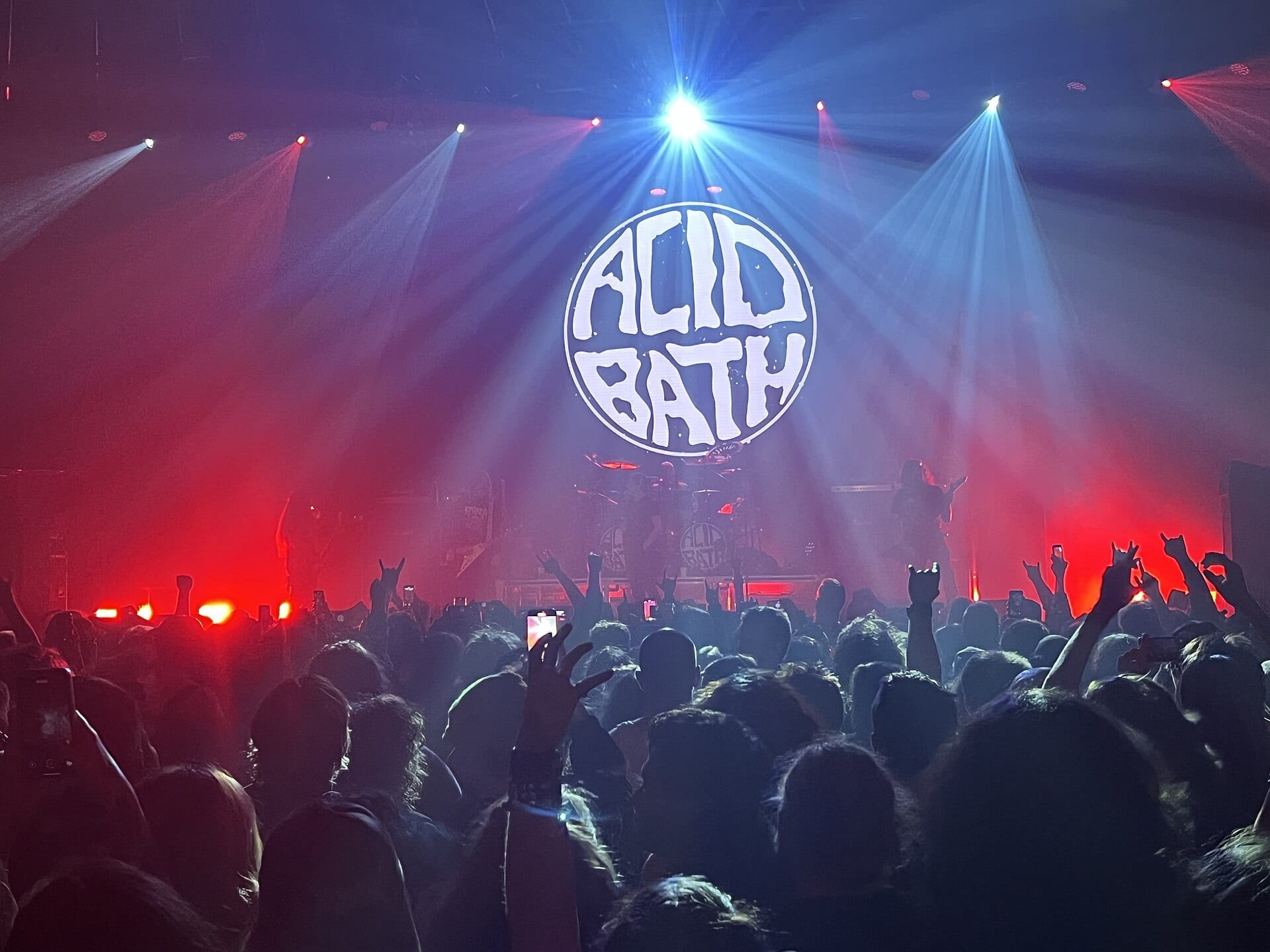 A club crowd throws up horns beneath red and blue beams, facing a stage backed by a circular white Acid Bath logo, the band lit only in silhouette.
