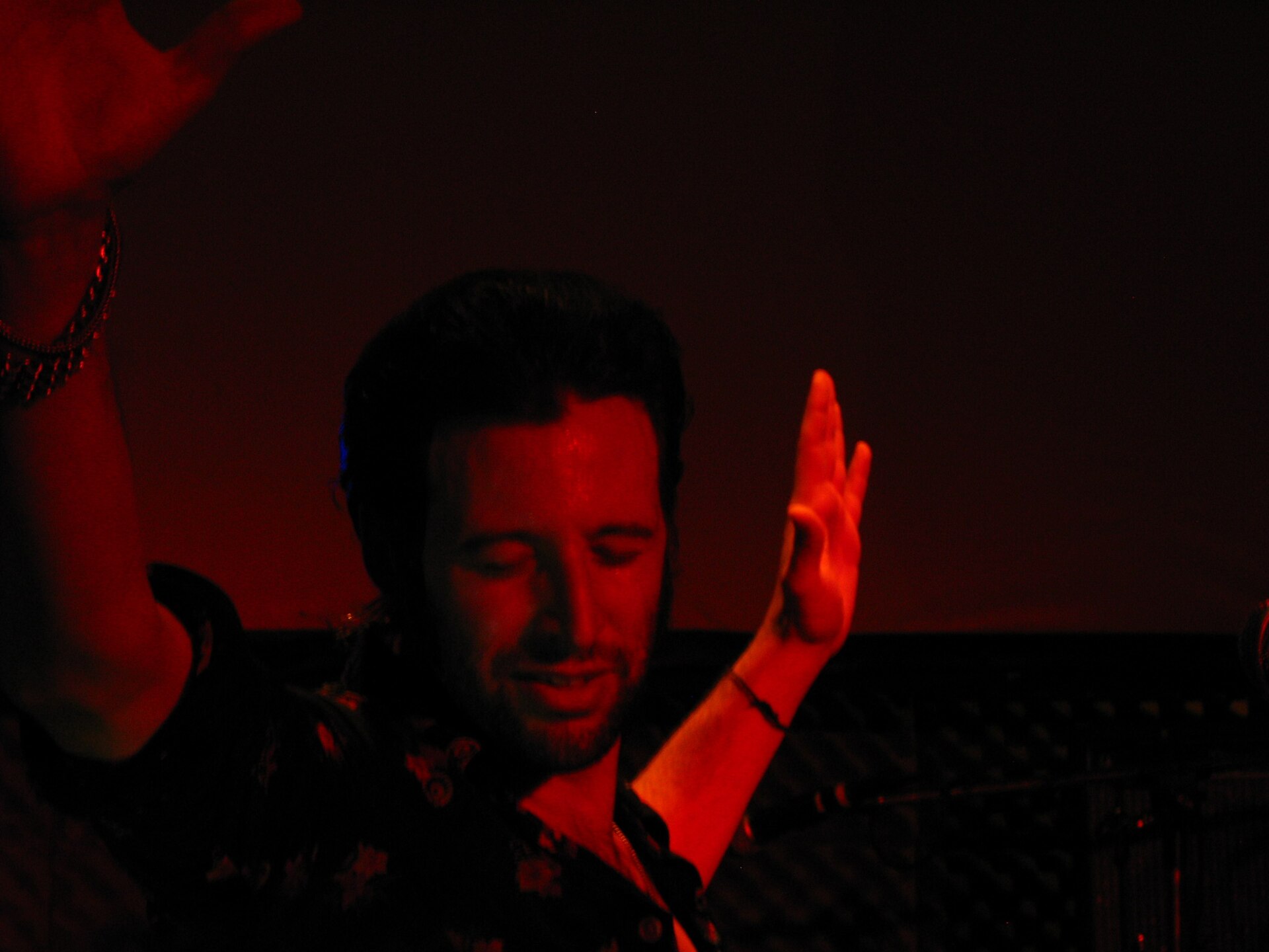 Adanowsky stands in red stage light with one hand raised, dark hair and a beard, a keyboard and cymbal beside him at the end of a set.