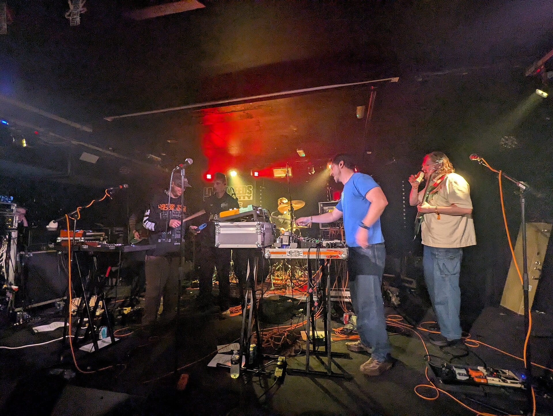Members of the band Adult DVD stand together on stage near amps and pedal boards under red stage lighting, one member singing into a handheld microphone.