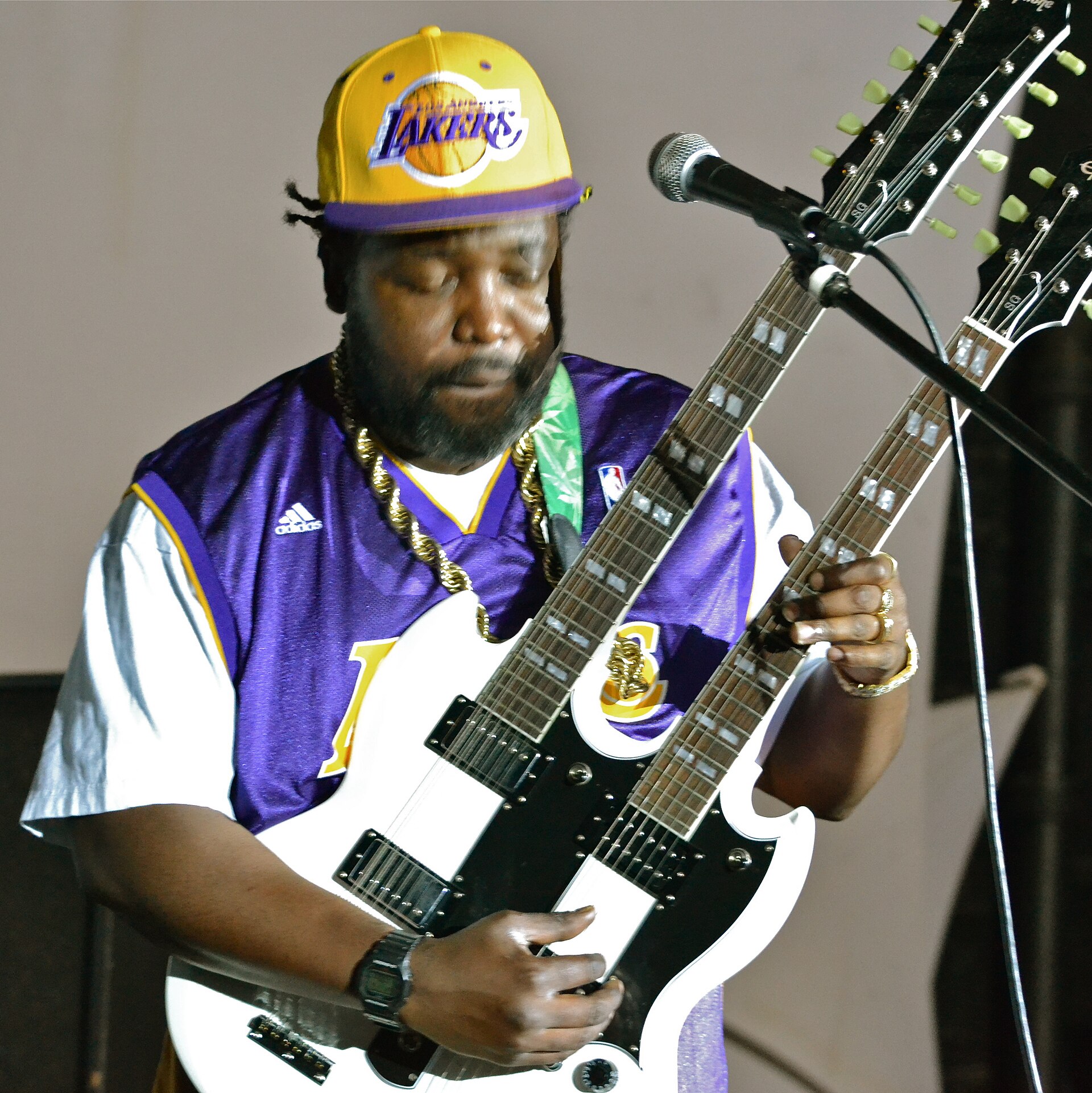 Afroman playing a white double-neck electric guitar on stage in a yellow Lakers cap, a purple Lakers jersey and a heavy gold rope chain