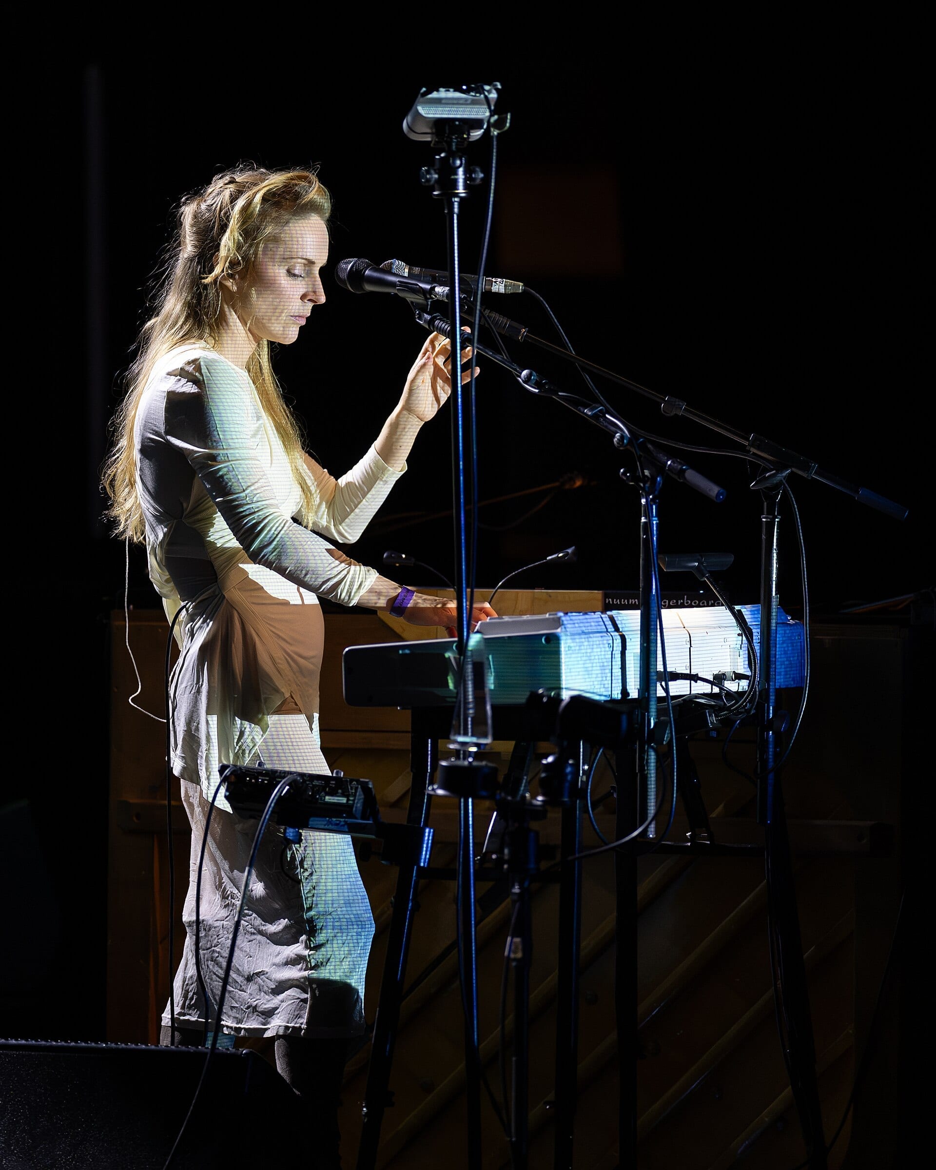 Agnes Obel stands in profile at a microphone behind a bank of keyboards, eyes closed, in a pale long-sleeved top with projected light falling across her.