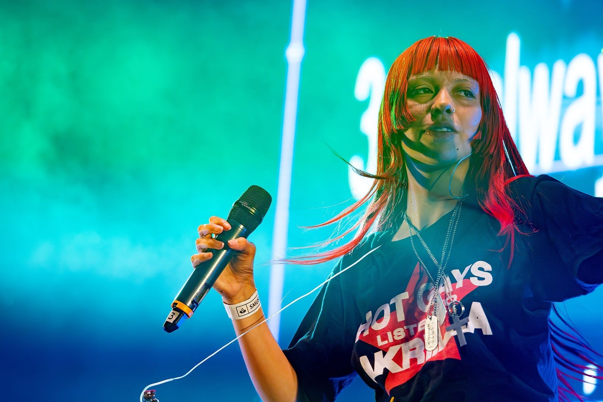 AKRIILA, with red-orange hair, sings into a handheld microphone on stage, wearing a black shirt that reads Hot Gays Listen to Akriila.