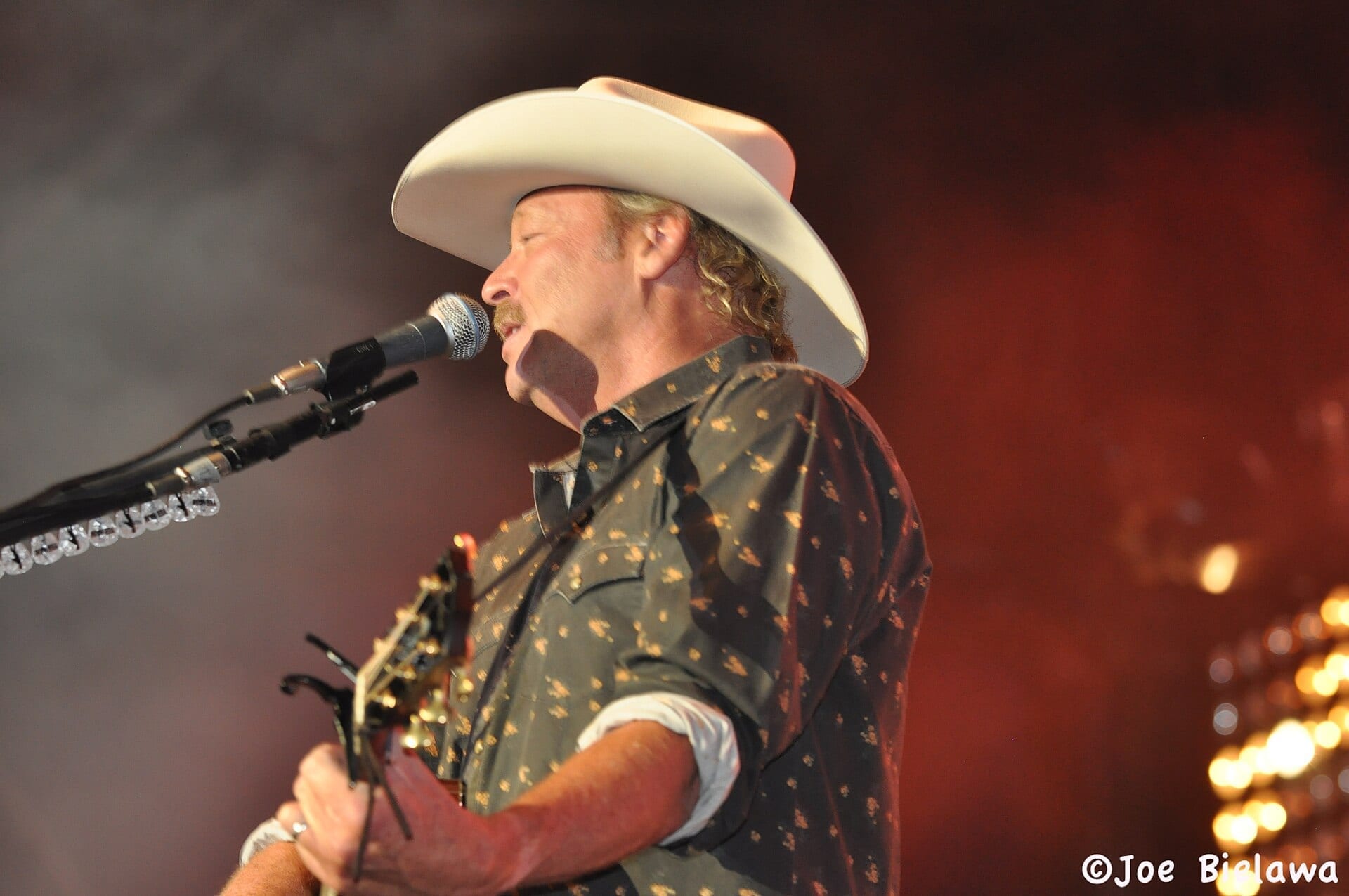 Alan Jackson sings in profile at a microphone in a white cowboy hat and a dark western shirt flecked with gold, one hand on a guitar neck.