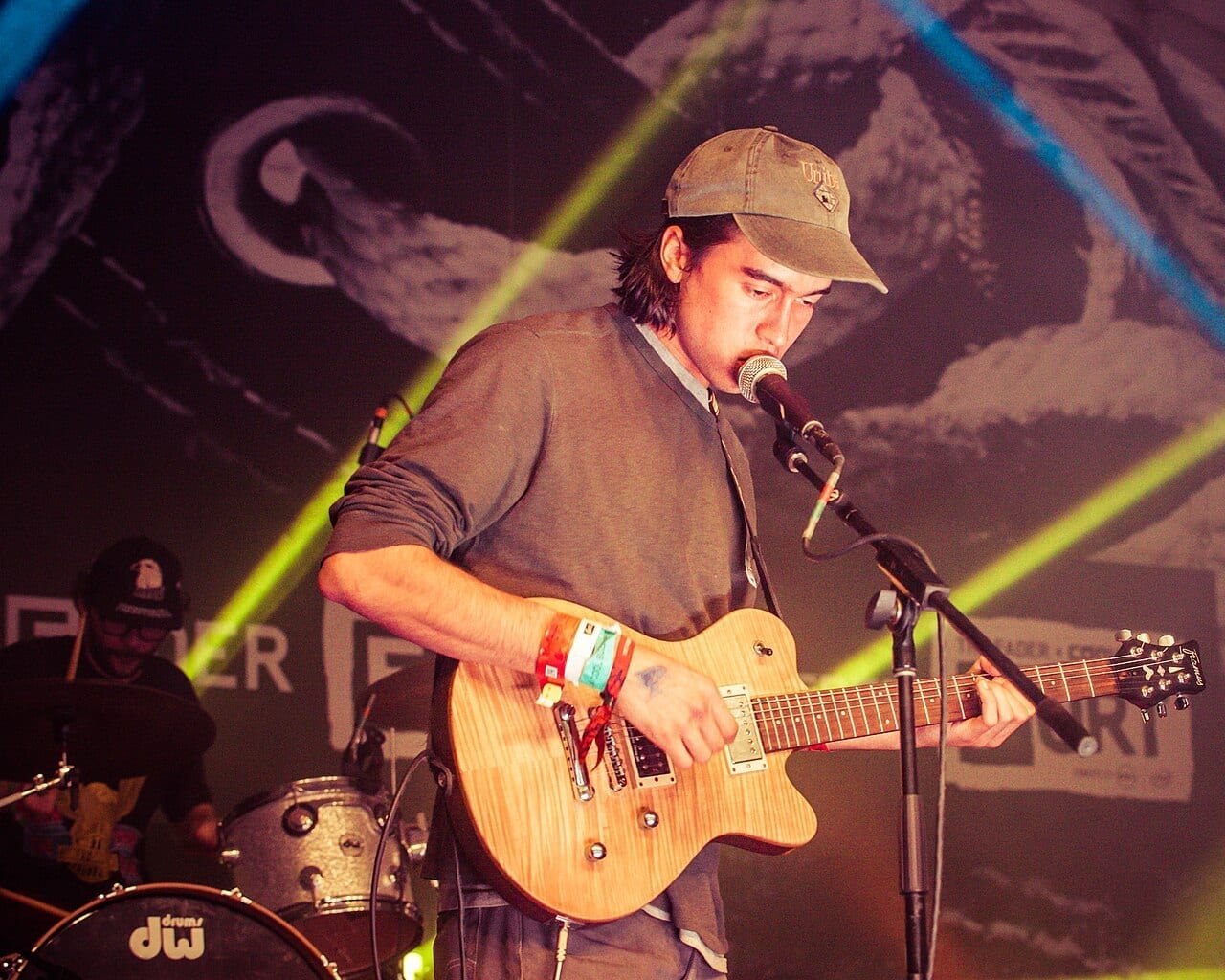 Alex G sings into a microphone while playing an electric guitar on stage, with a drummer behind him.