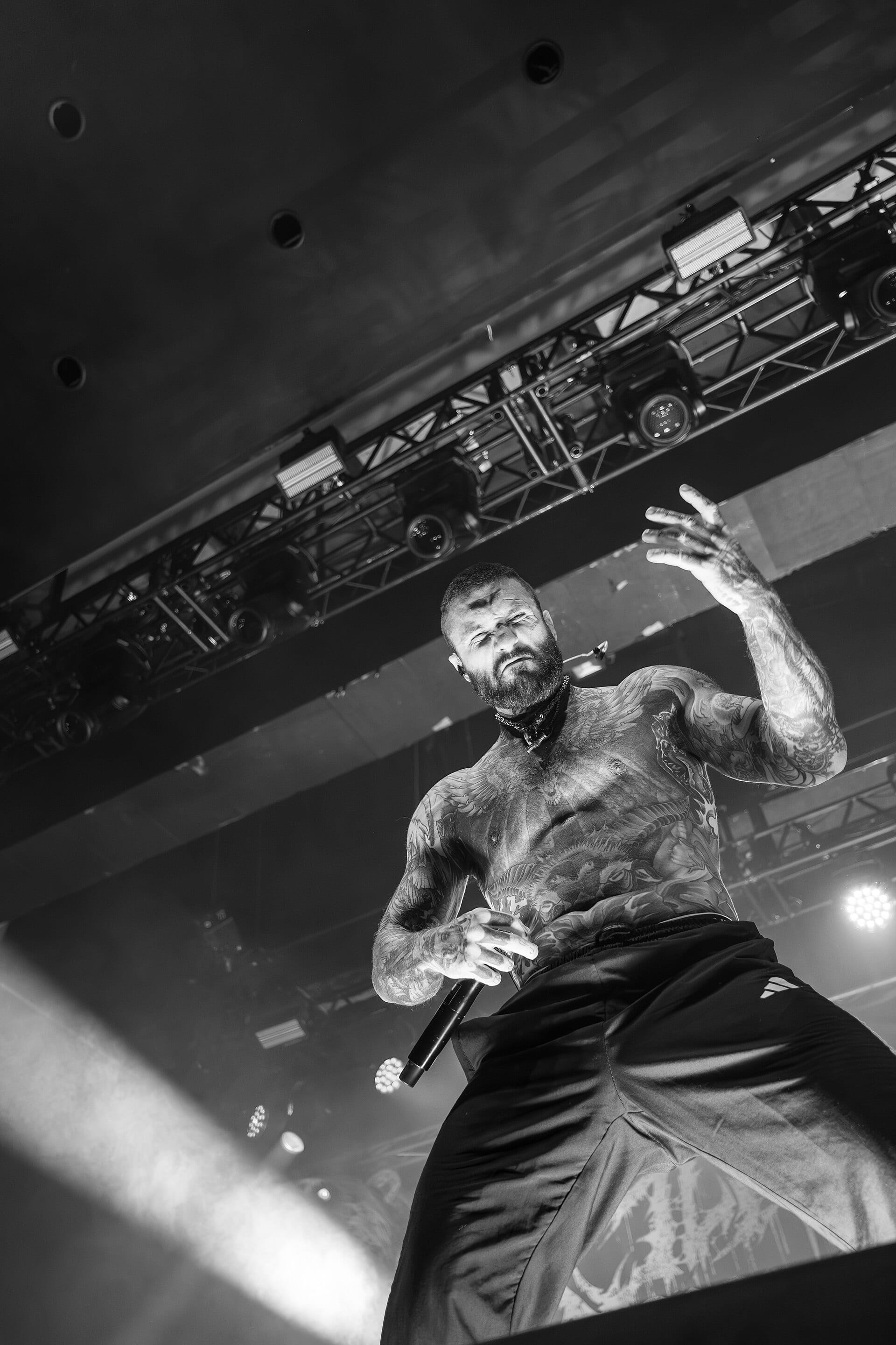 Alex Terrible performs shirtless and heavily tattooed, gripping a microphone and raising one hand, photographed from below the stage in black and white.
