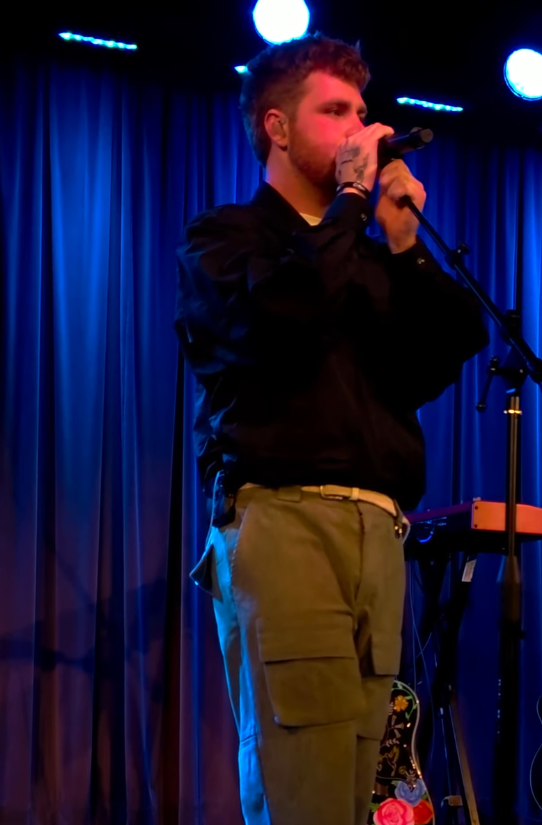 Alex Warren sings into a microphone on a stand in a black jacket and olive cargo trousers, blue curtain behind him.