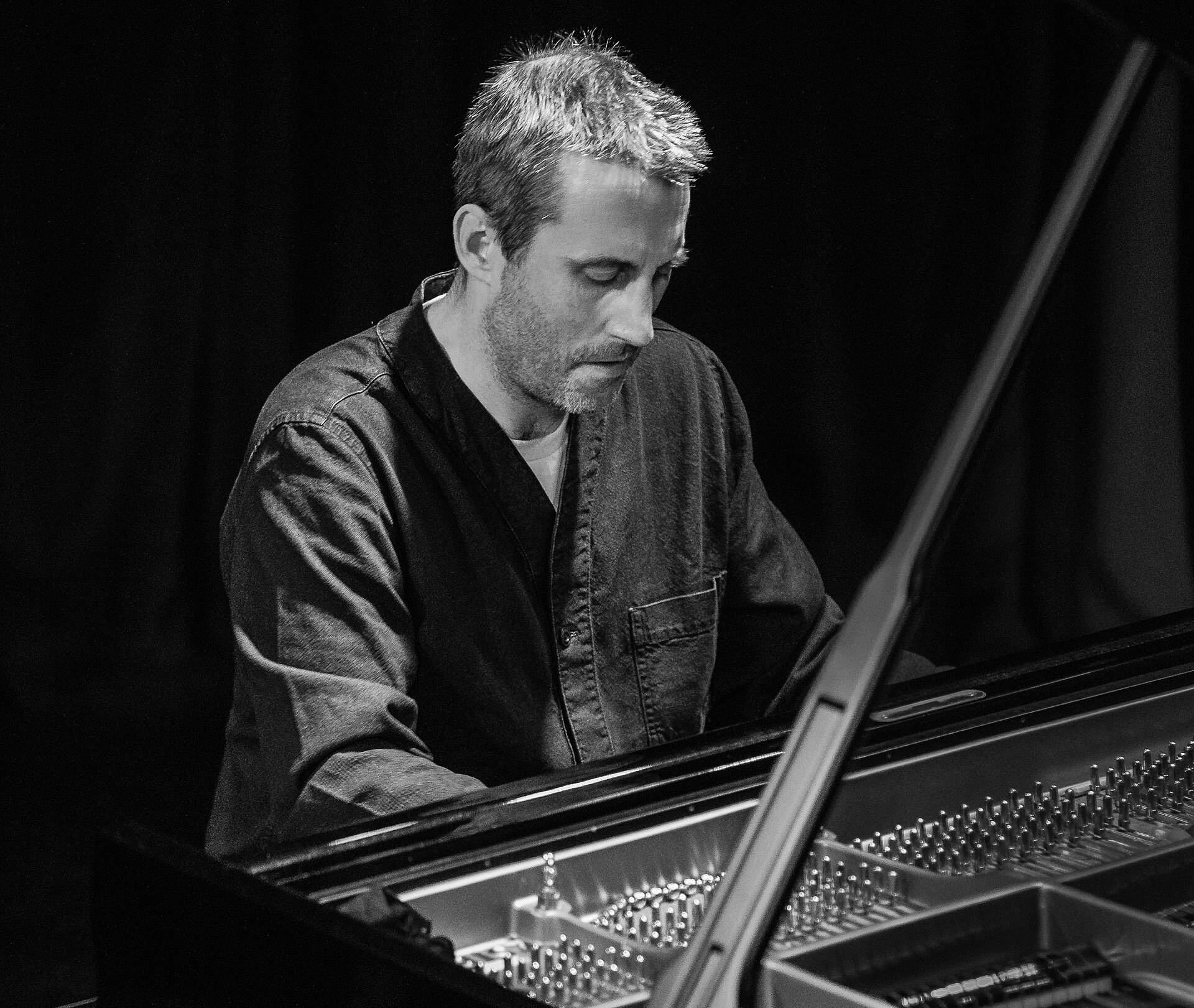 A pianist in a dark work jacket looks down at the keys of a grand piano, photographed in black and white against a dark stage