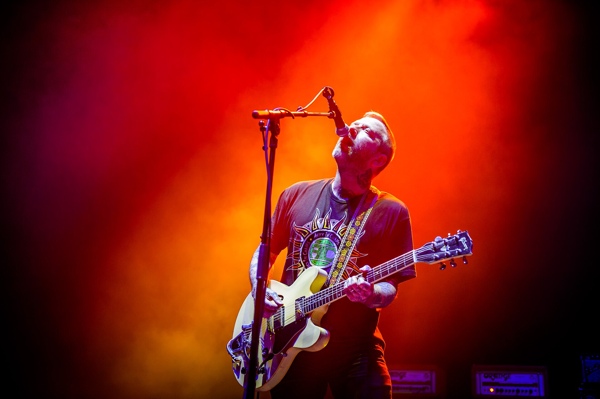 An Alexisonfire guitarist throws his head back to sing into an overhead microphone, playing a cream semi-hollow guitar against a wall of orange and red stage haze.