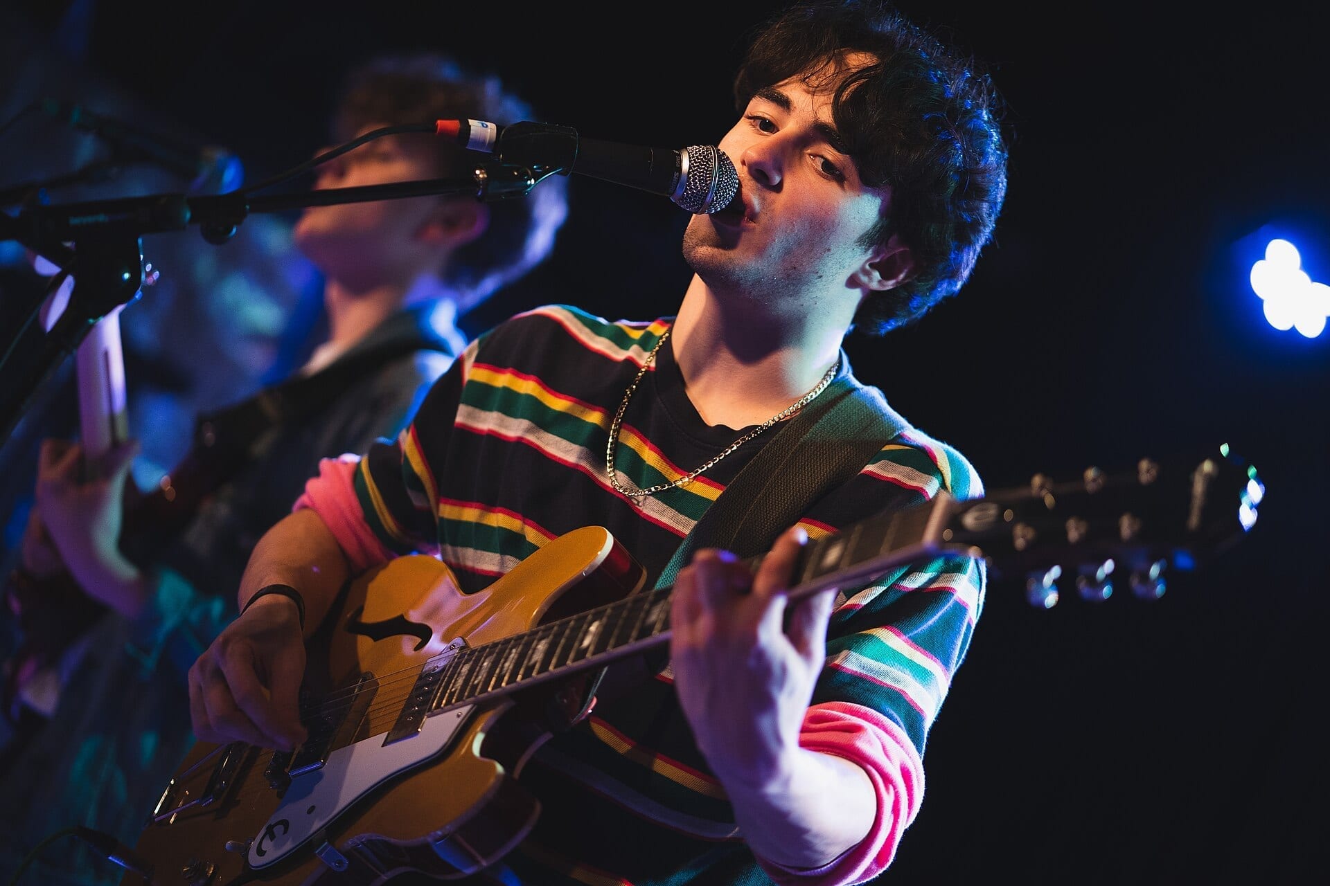 Alfie Templeman sings into a microphone under blue and pink stage lighting while playing a mustard-yellow semi-hollow electric guitar, wearing a striped shirt and a gold chain, with a bandmate playing guitar in the background.