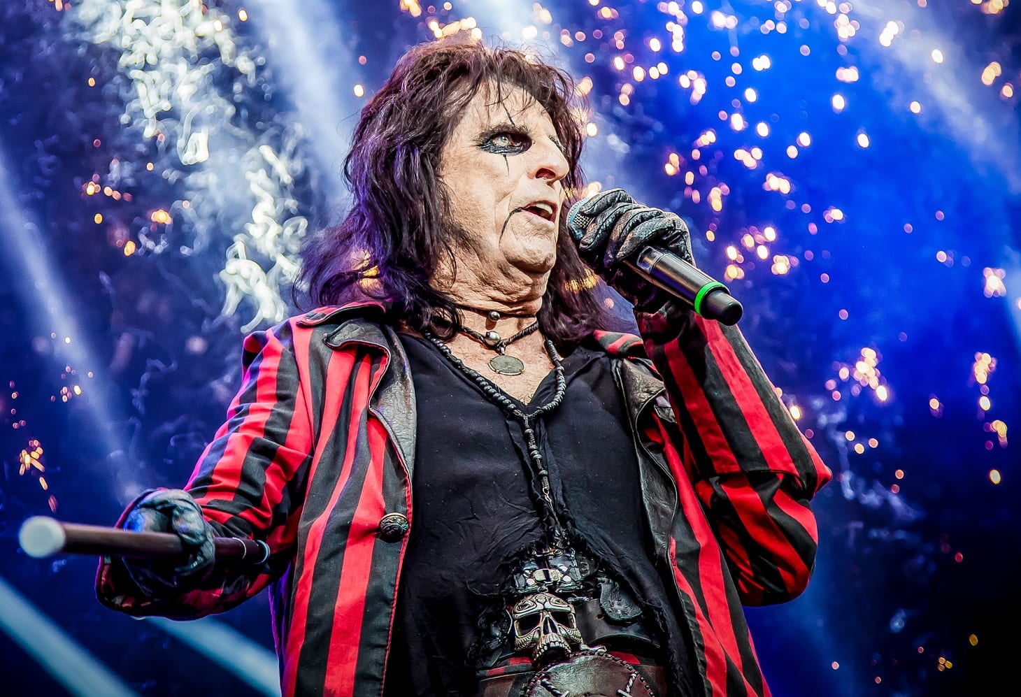 Alice Cooper sings into a microphone in heavy black eye make-up and a red and black striped jacket, sparks falling behind him.
