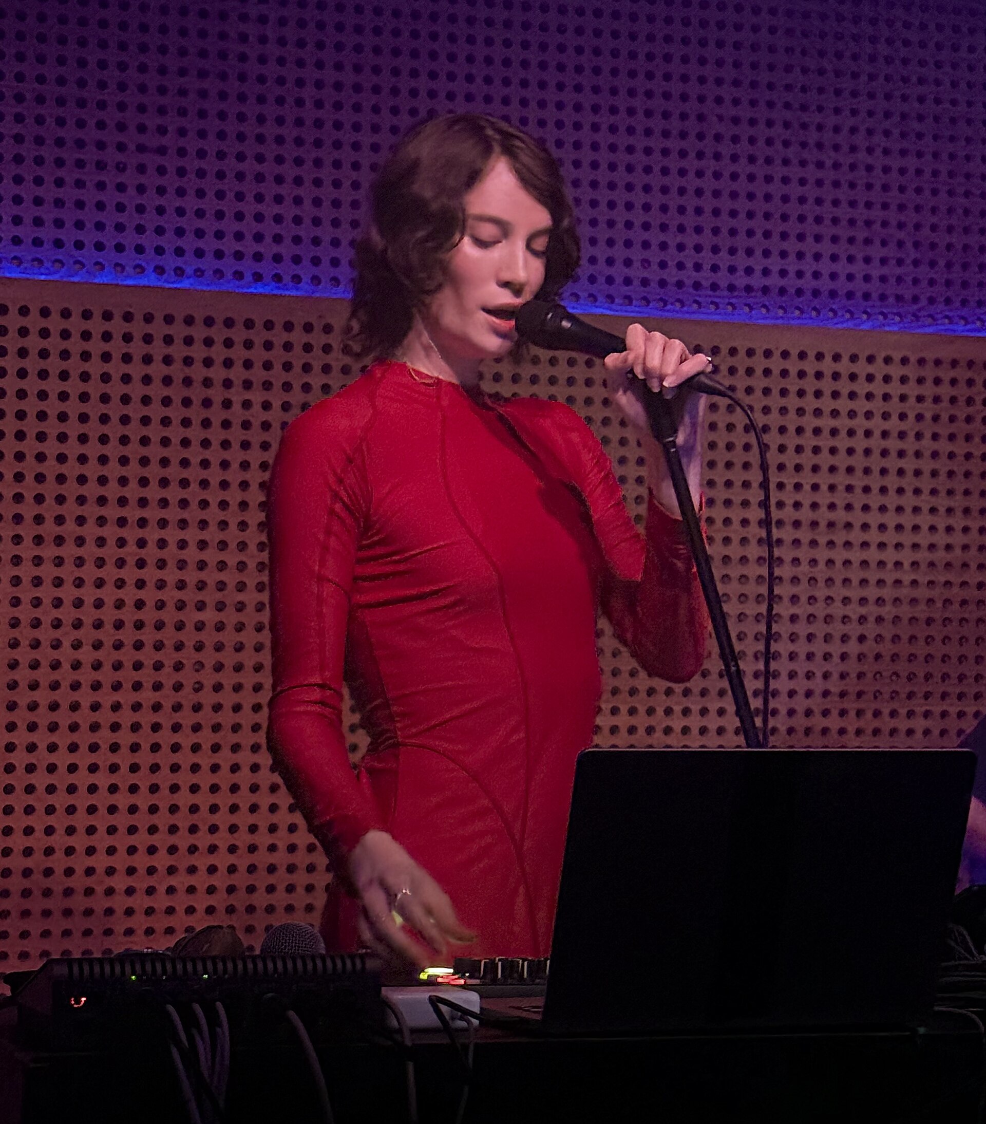 Alif Hilal sings into a handheld microphone behind a laptop and a mixer, wearing a long-sleeved red dress, against a perforated wall lit red and blue.