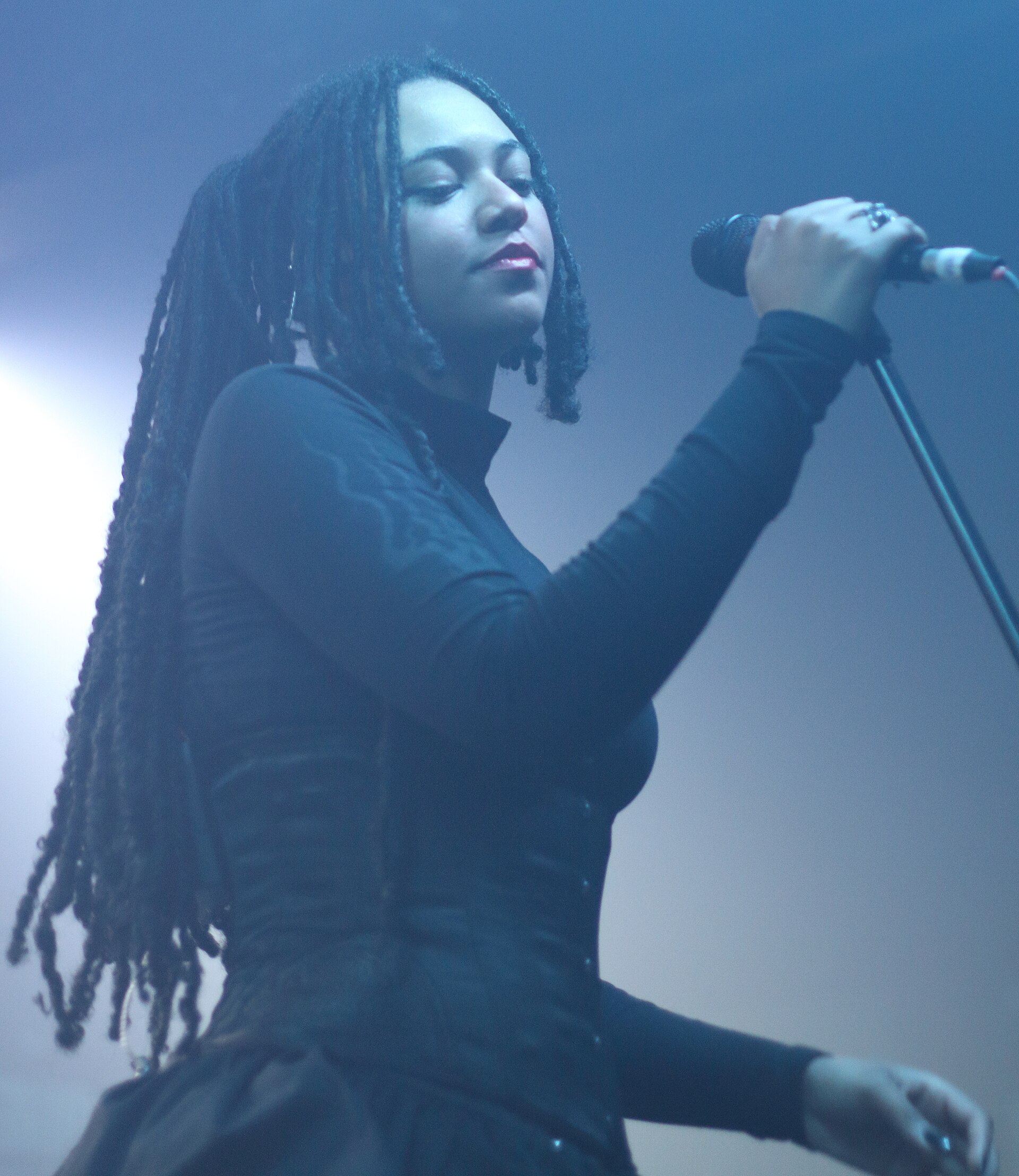 A singer with long locs holds a microphone on its stand under cold blue light, in a black long-sleeved top.