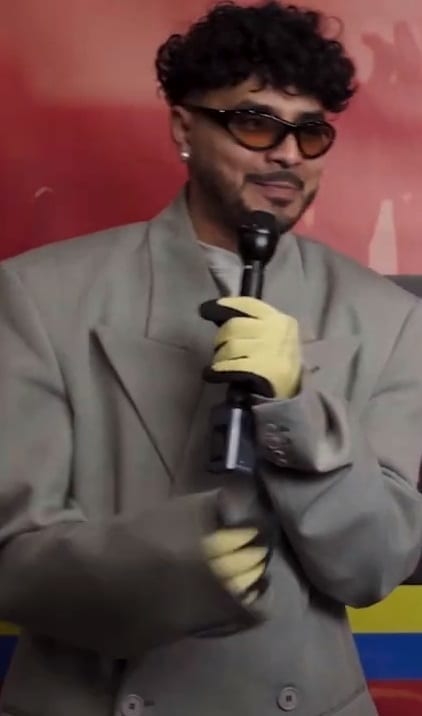 Alvaro Diaz, wearing tinted sunglasses and an oversized grey suit with yellow gloves, holds a microphone during an interview.