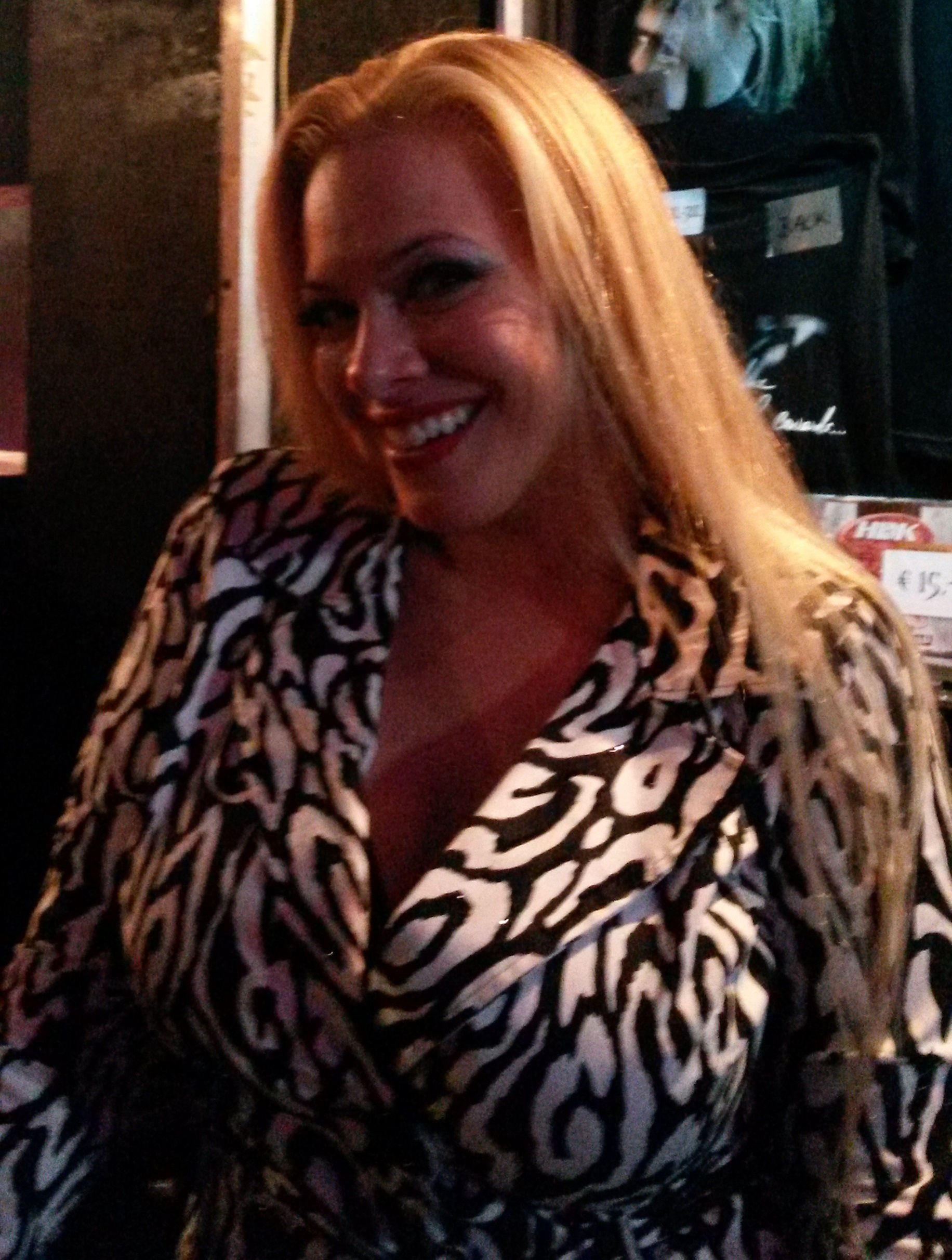 A smiling woman with long blonde hair in a patterned wrap top, photographed close up in a dimly lit venue