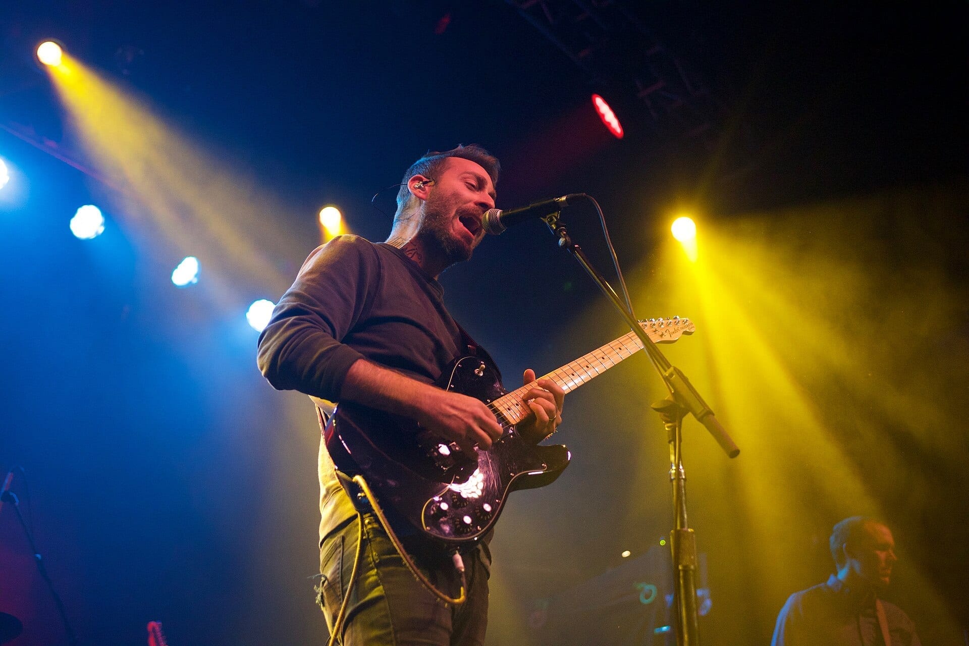 Mike Kinsella of American Football sings into a microphone while playing a guitar, lit by blue and yellow stage lights.