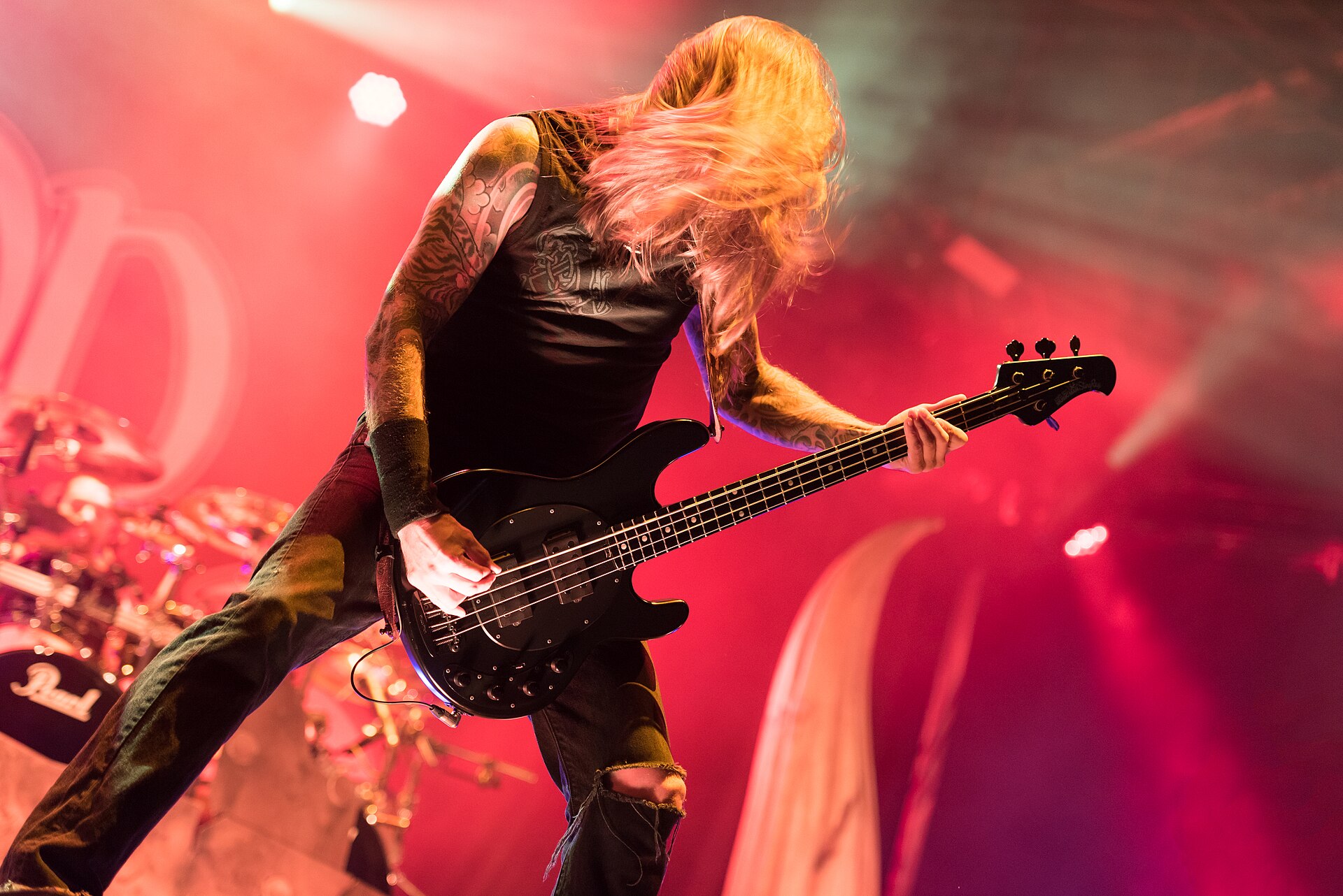 A long-haired, heavily tattooed bassist swings his hair forward mid-song under deep red stage light.
