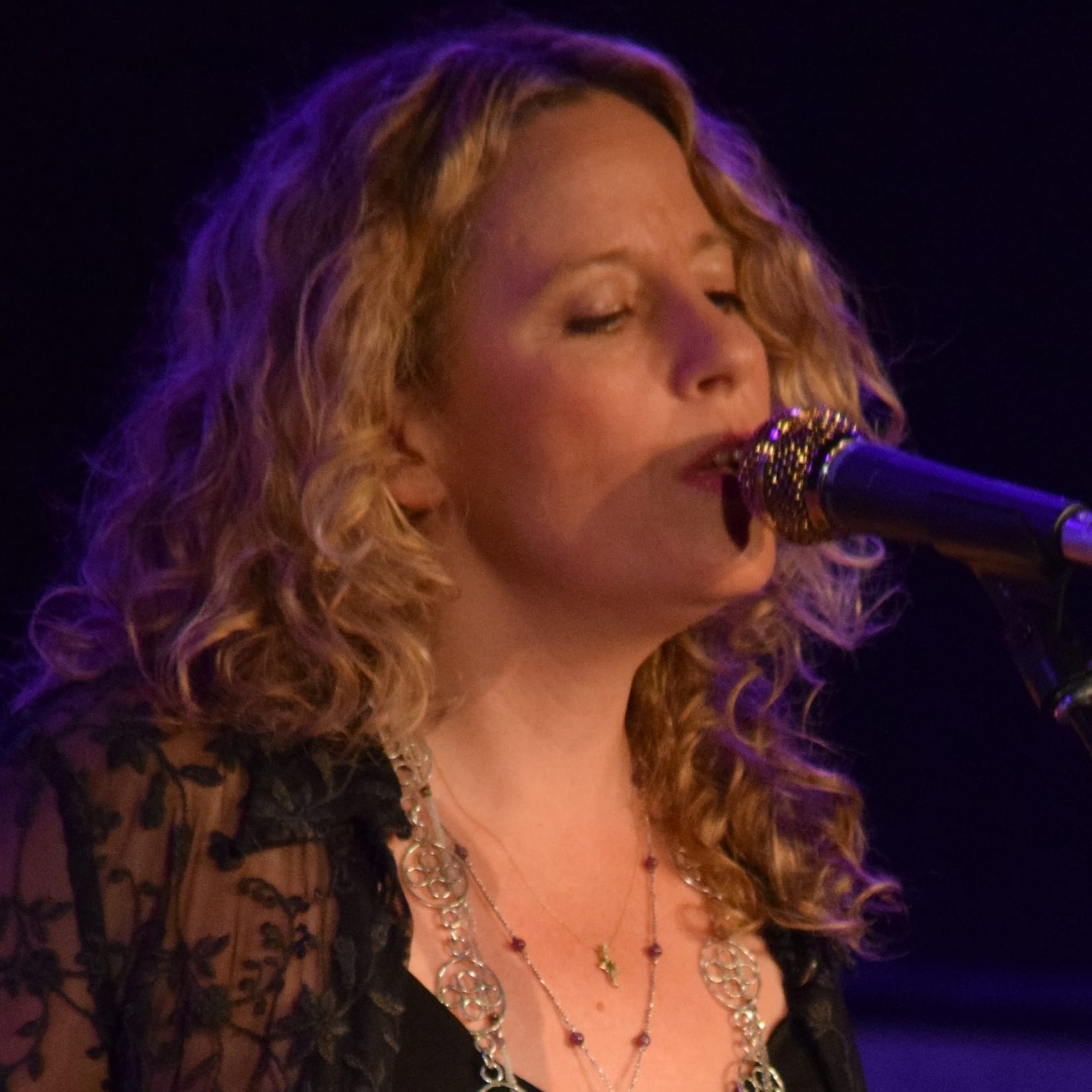 Amy Helm sings into a microphone with her eyes closed, curly blonde hair falling around her face, wearing layered necklaces under purple stage lighting.
