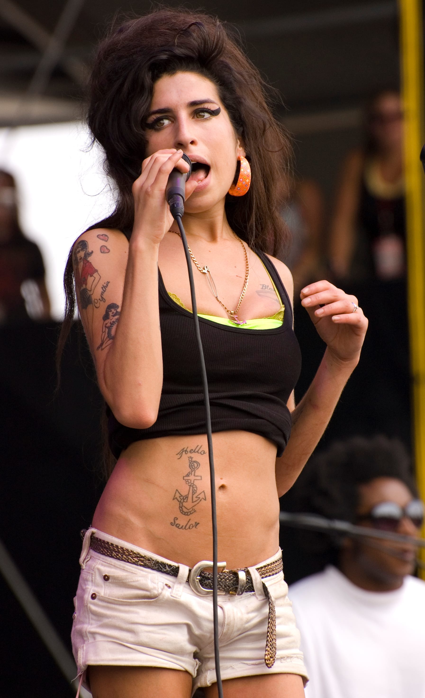Amy Winehouse sings into a handheld microphone in a black vest and white shorts, tattoos down her arm and her hair piled into a beehive.