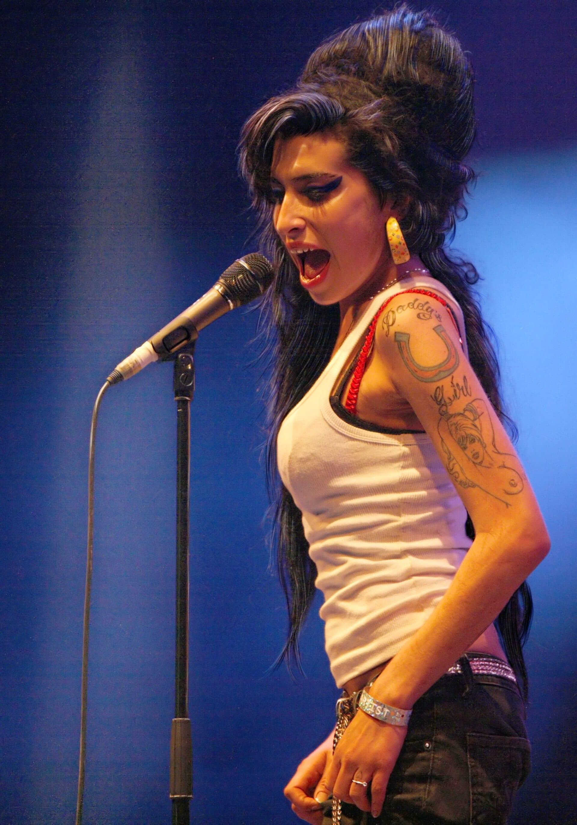 Amy Winehouse sings open-mouthed at a microphone stand in profile, her beehive piled high, wearing a white vest that shows the horseshoe tattoo on her upper arm.