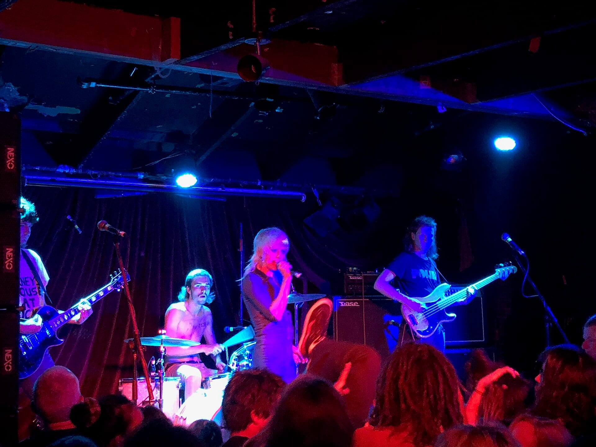 Amyl and the Sniffers perform on a small club stage lit in blue and red, the singer holding a microphone between two guitarists and a shirtless drummer, in front of a packed crowd.