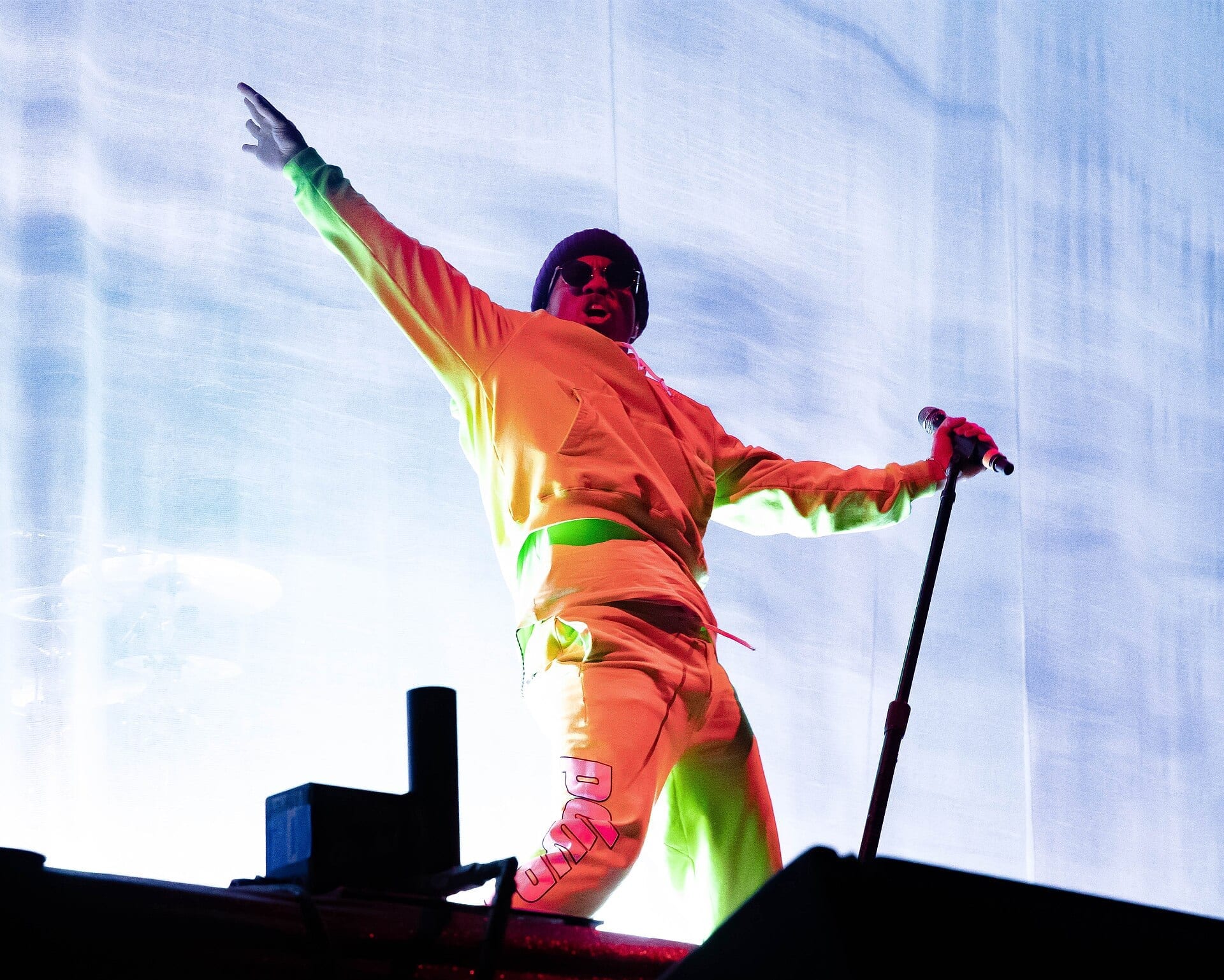 Anderson .Paak throws one arm out while holding a mic stand with the other, in an orange tracksuit and sunglasses, lit green and pink against a white backdrop.