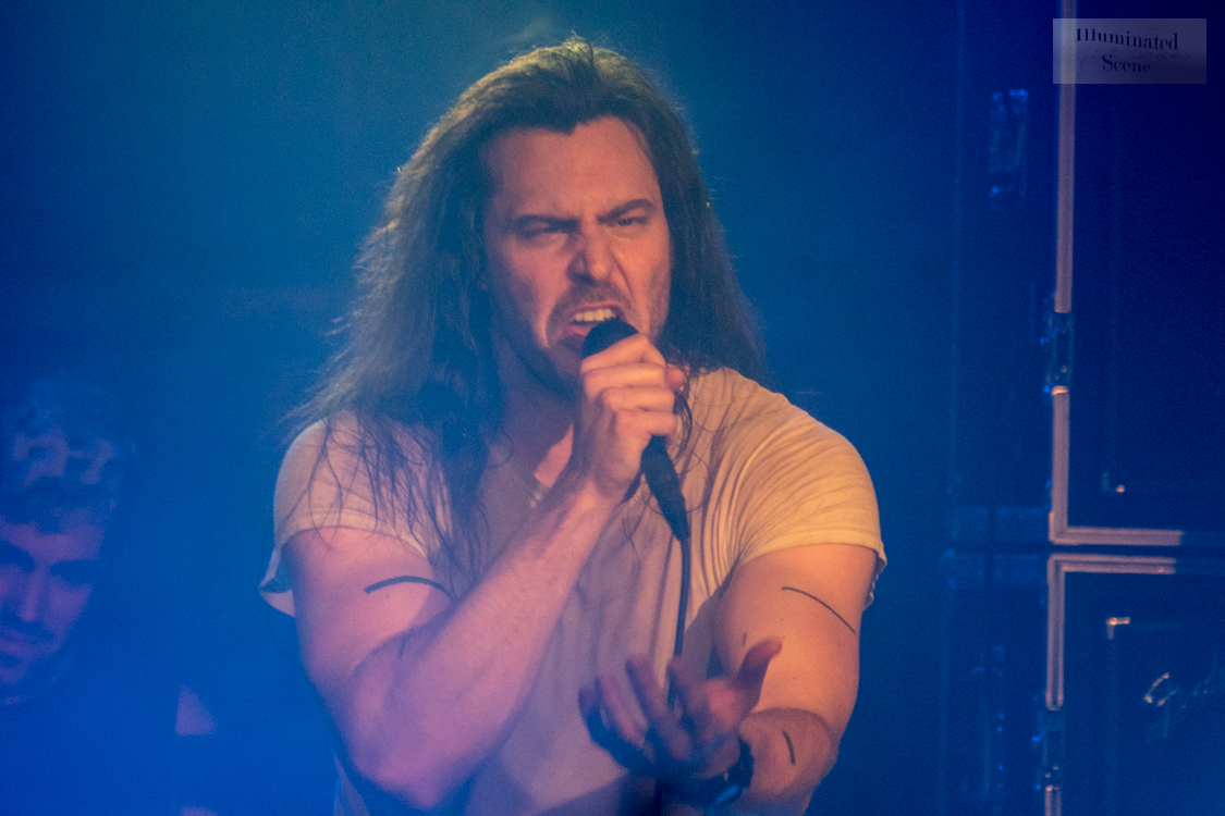 Andrew W.K. leans back with his eyes shut and shouts into a handheld microphone, long dark hair falling across his face, in a white shirt.
