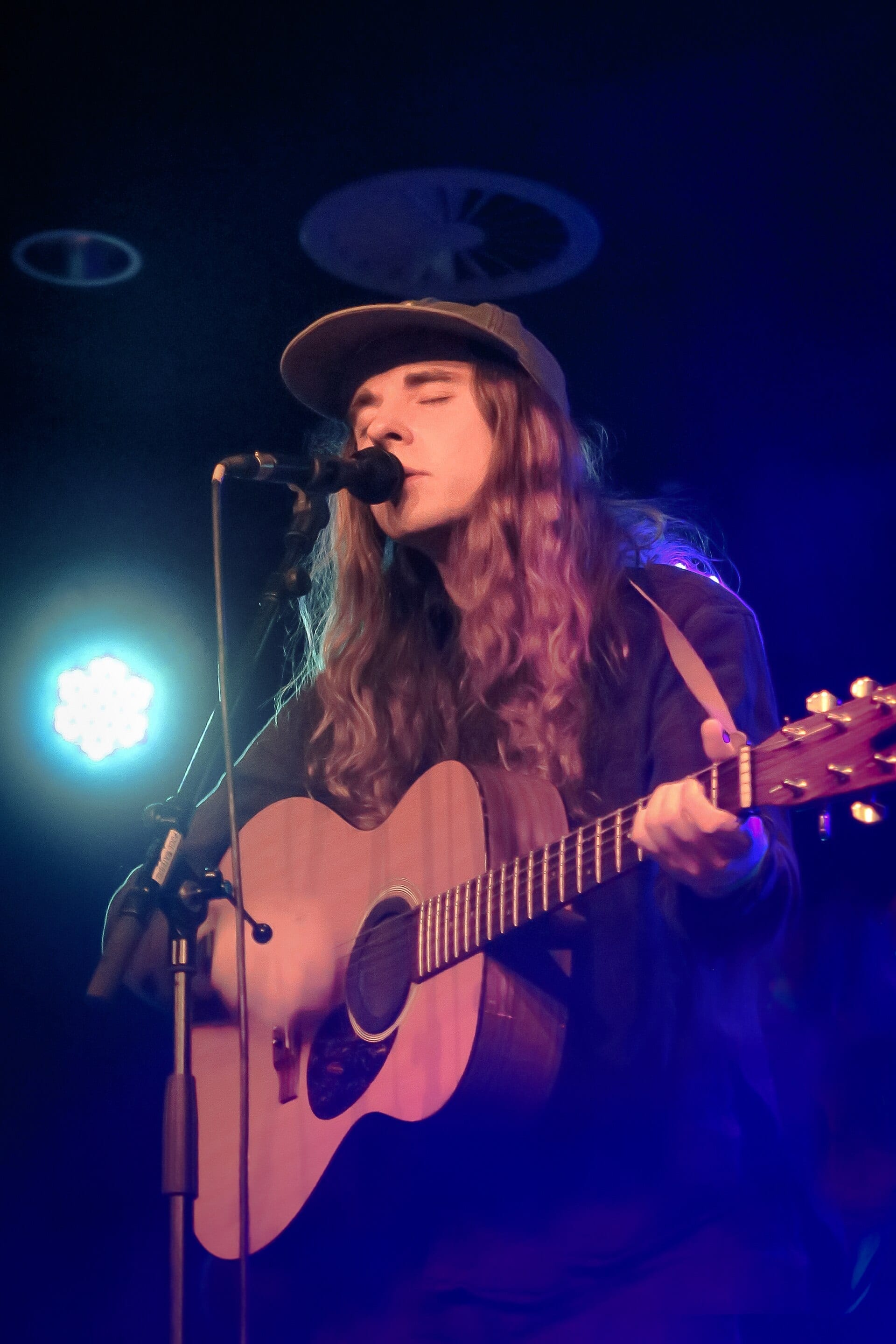Andy Shauf, with long curly hair under a ball cap and his eyes closed, sings into a microphone while playing an acoustic guitar under blue stage light.