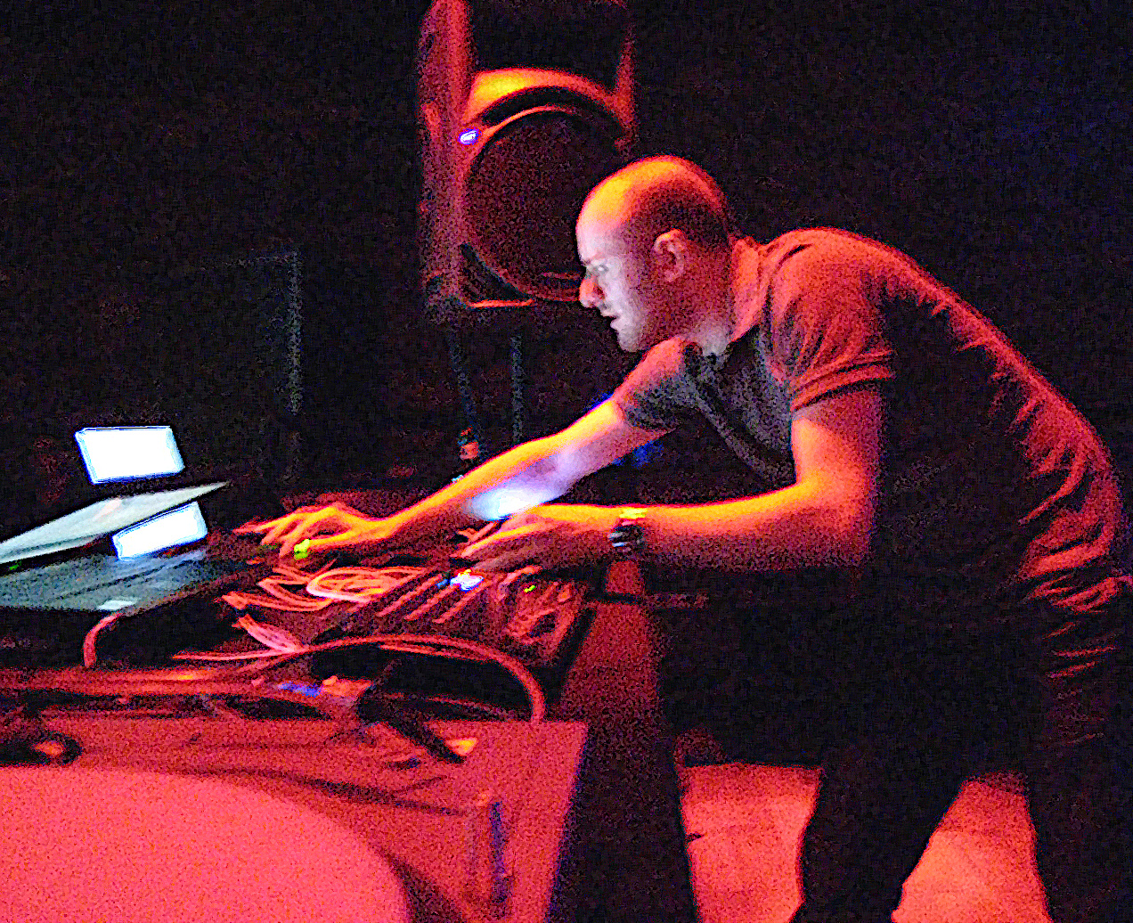 Andy Stott leans over a table of mixers and cables in red light, both hands on the controls, an open laptop to his left.