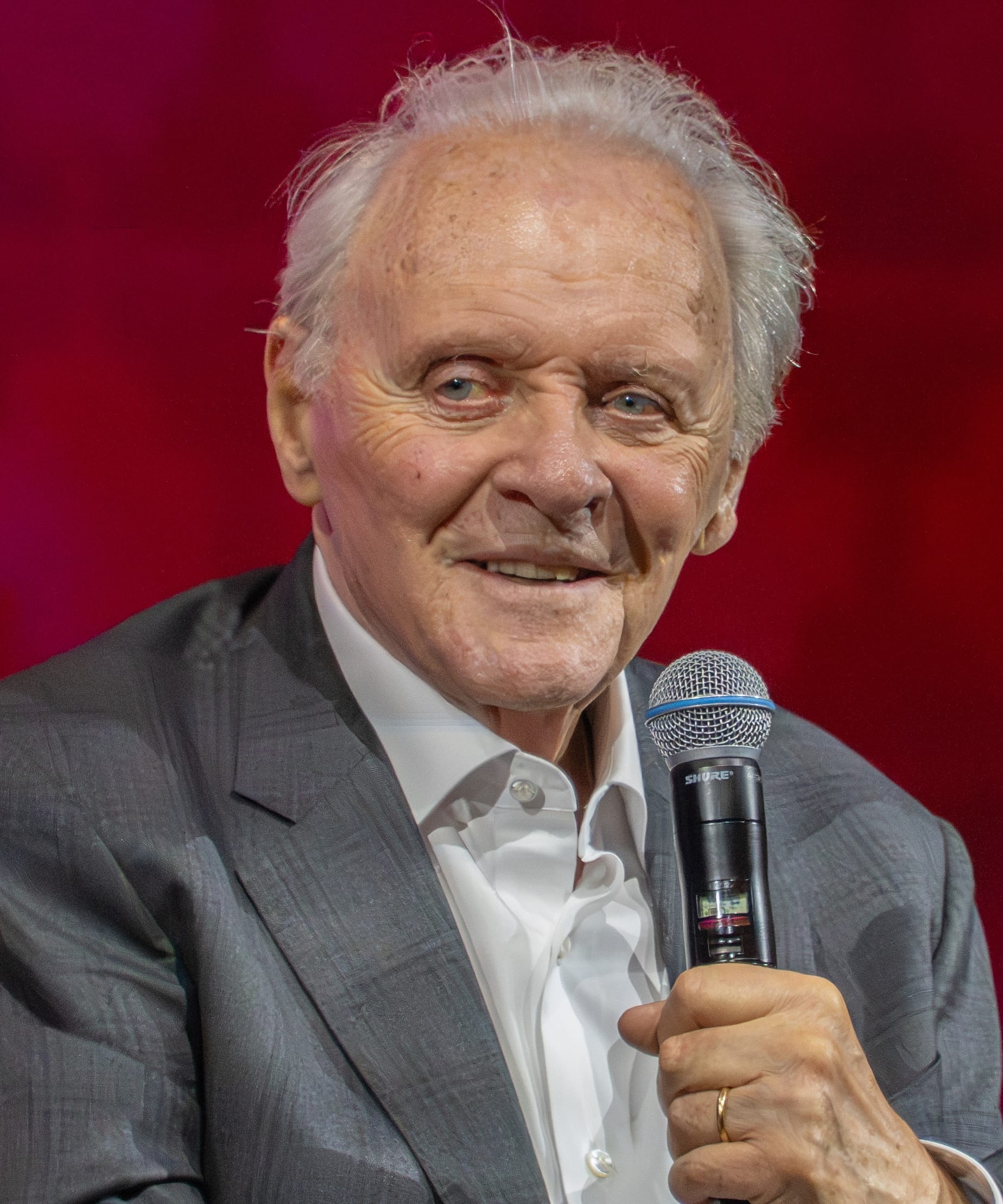 Anthony Hopkins holds a handheld microphone and smiles in a grey suit and open white shirt against a red backdrop.