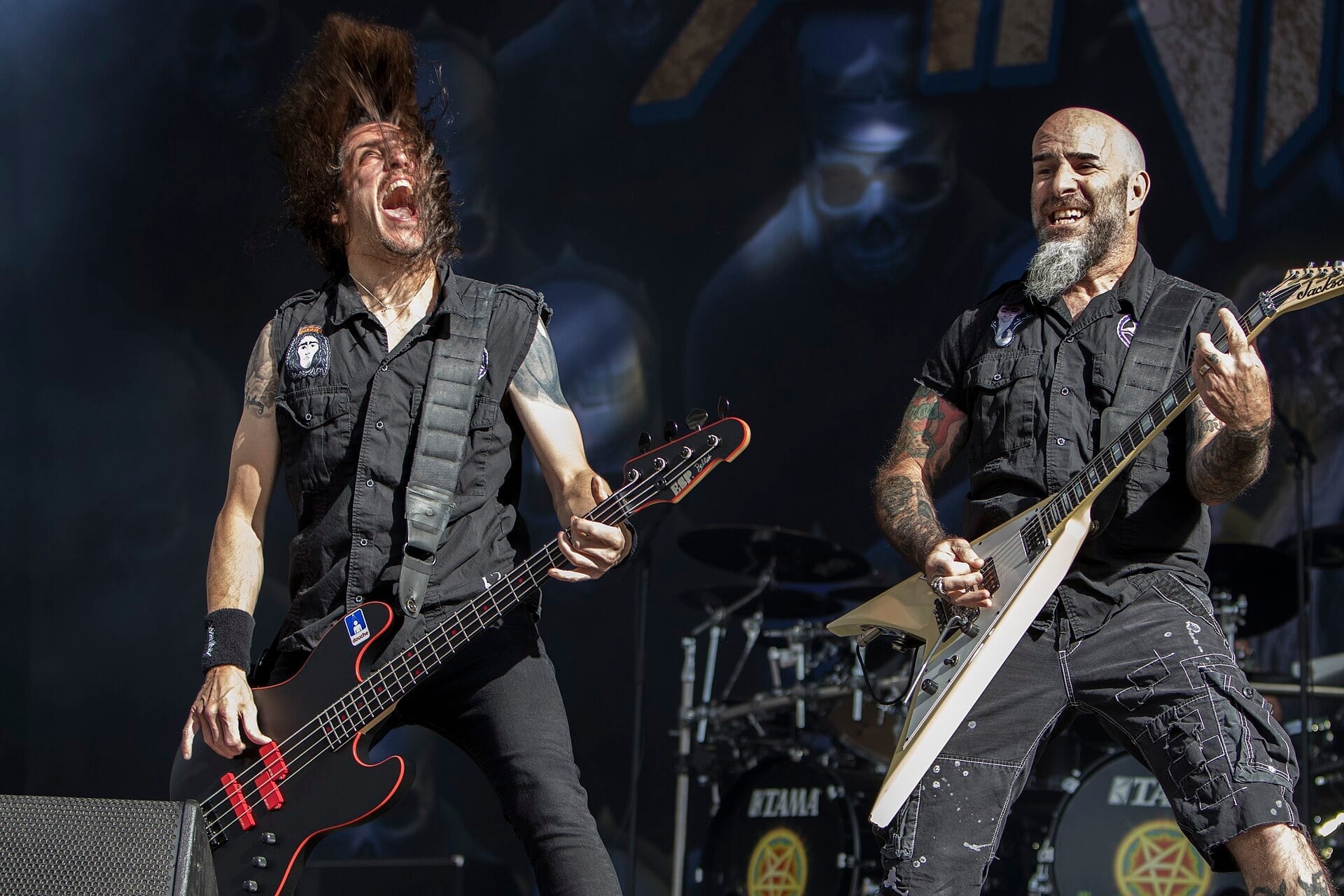 Two members of Anthrax perform live, one headbanging while playing a bass guitar and the other playing a white V-shaped guitar, with the band's pentagram logo visible behind them.