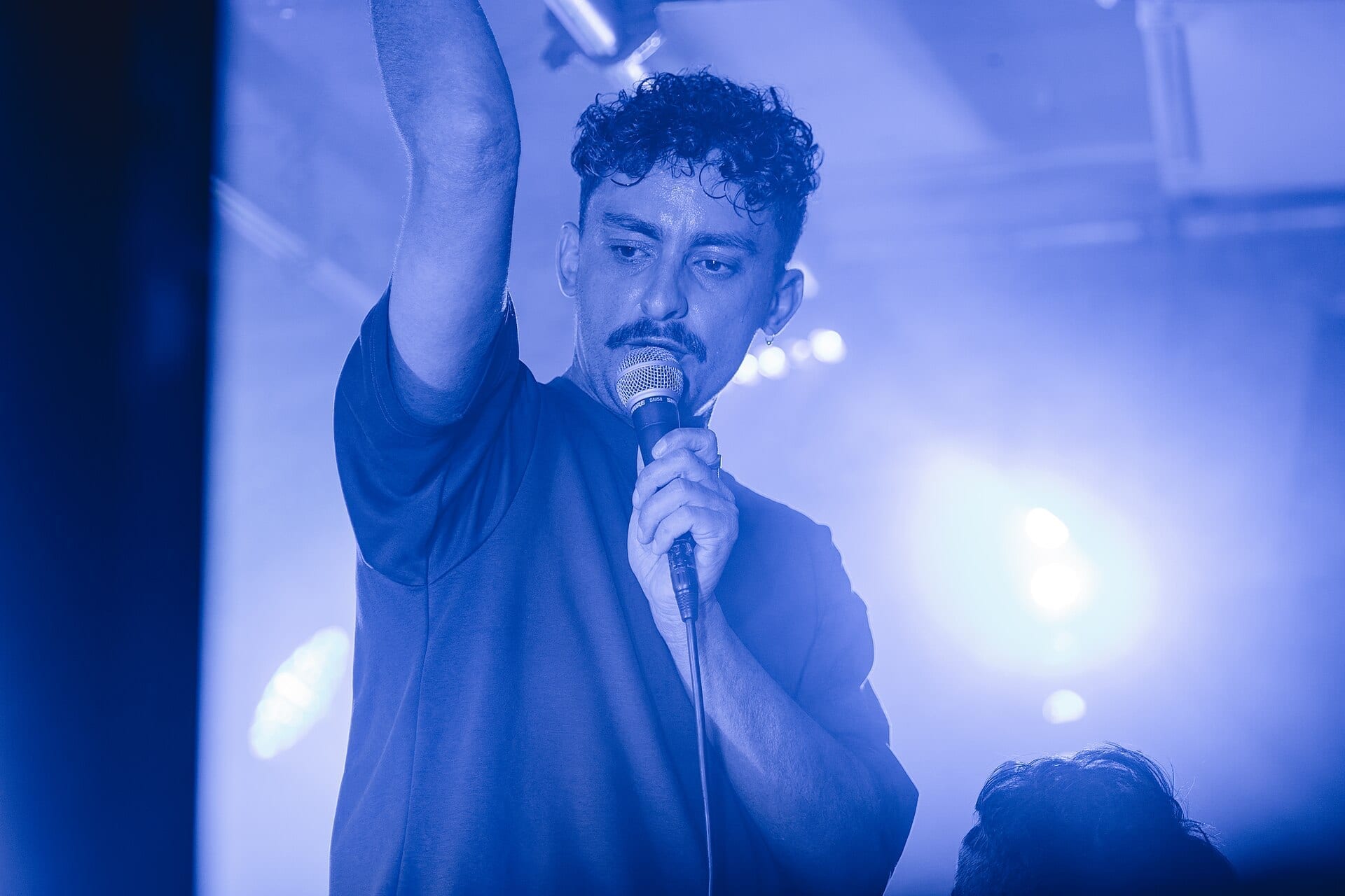 Antony Szmierek sings into a microphone with one arm raised, sporting a mustache and curly hair, under blue stage lighting.
