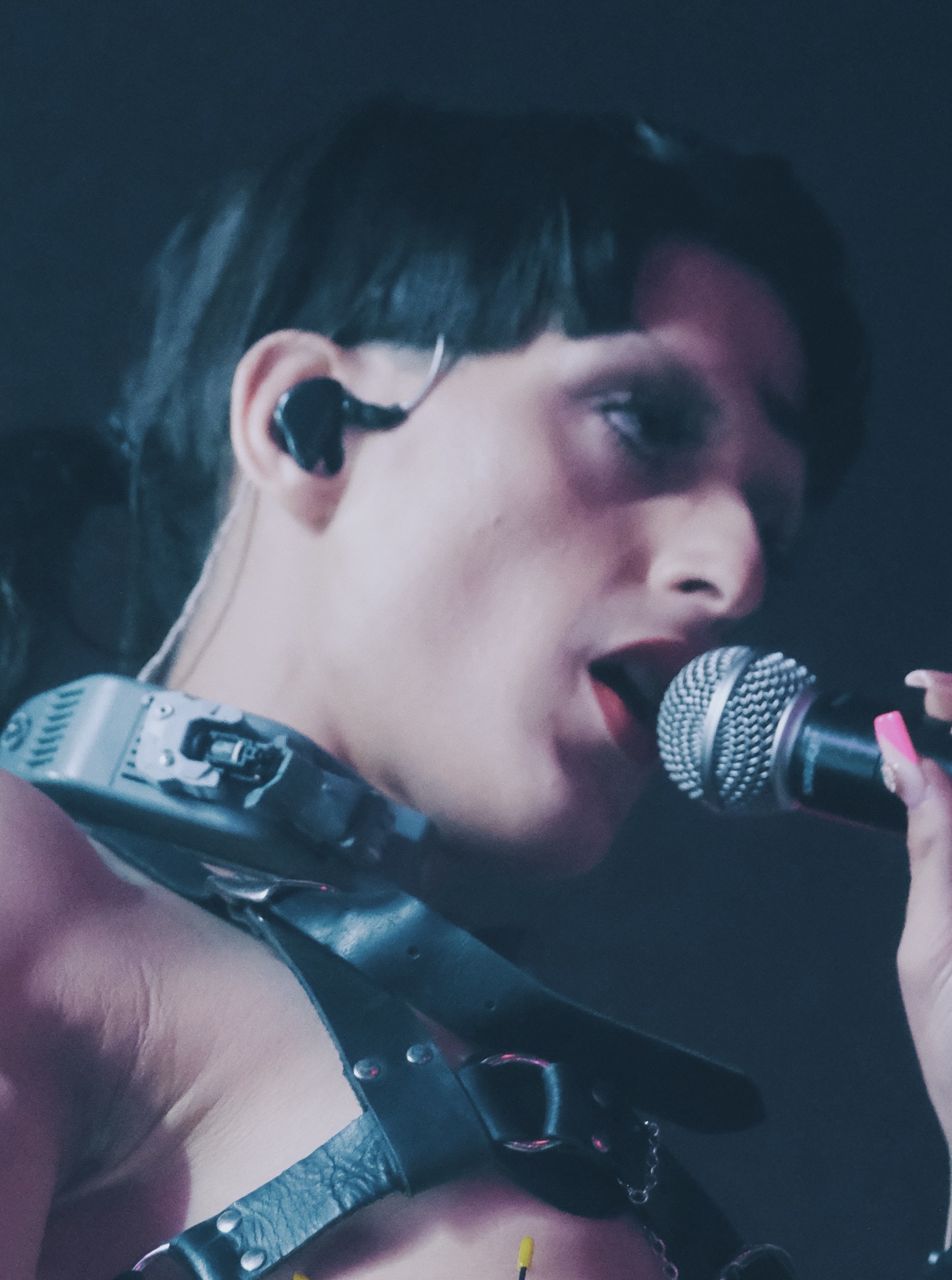 A performer in a studded leather harness and dark eye make-up sings into a handheld microphone, shot close and tightly cropped in low blue light