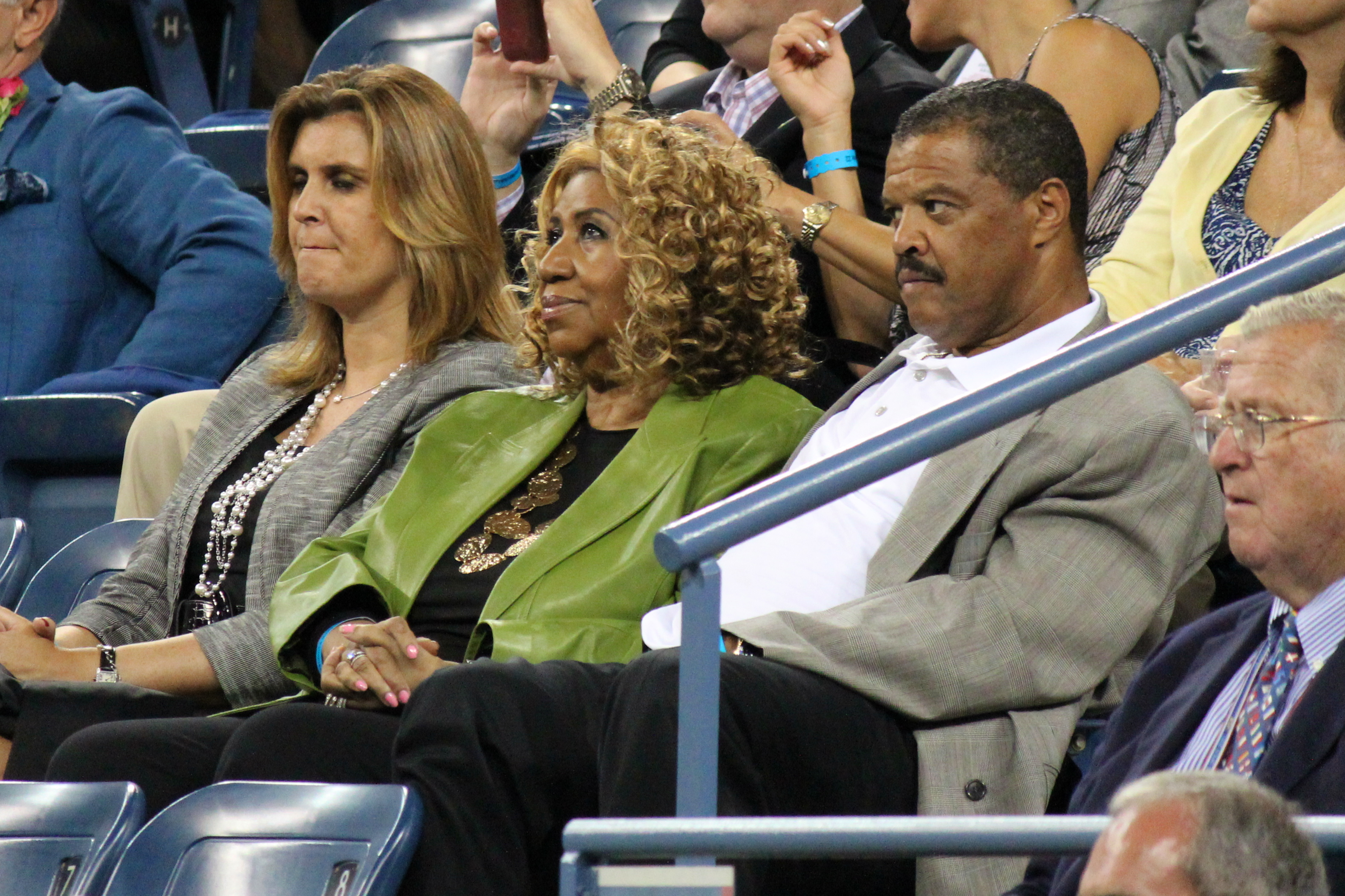 Aretha Franklin sits in a stadium seat in a green jacket over a black top, watching the court, with other spectators either side of her.