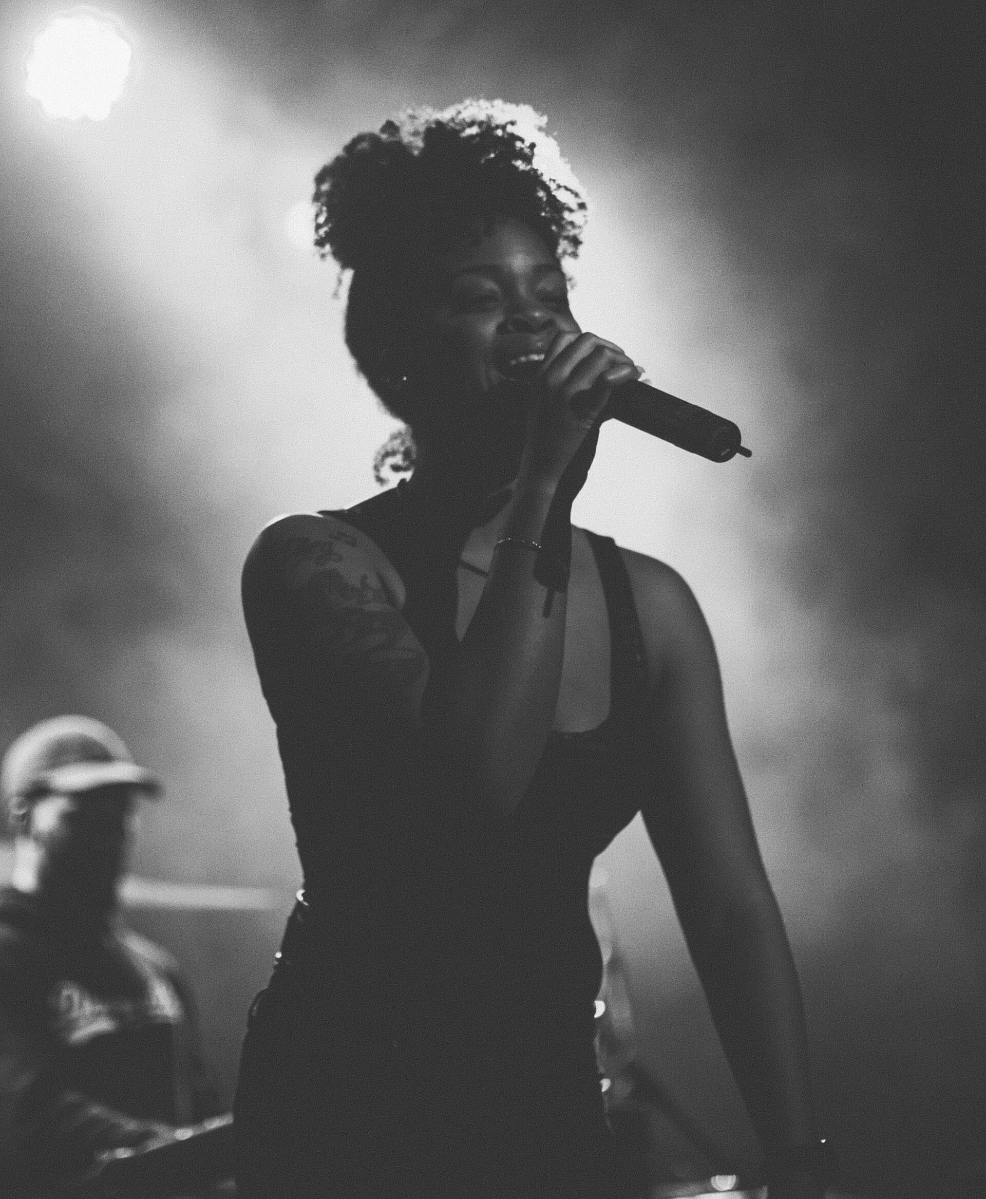 Ari Lennox sings into a microphone on a smoky, backlit stage, her curly hair silhouetted against bright stage lights, in a black and white photograph.