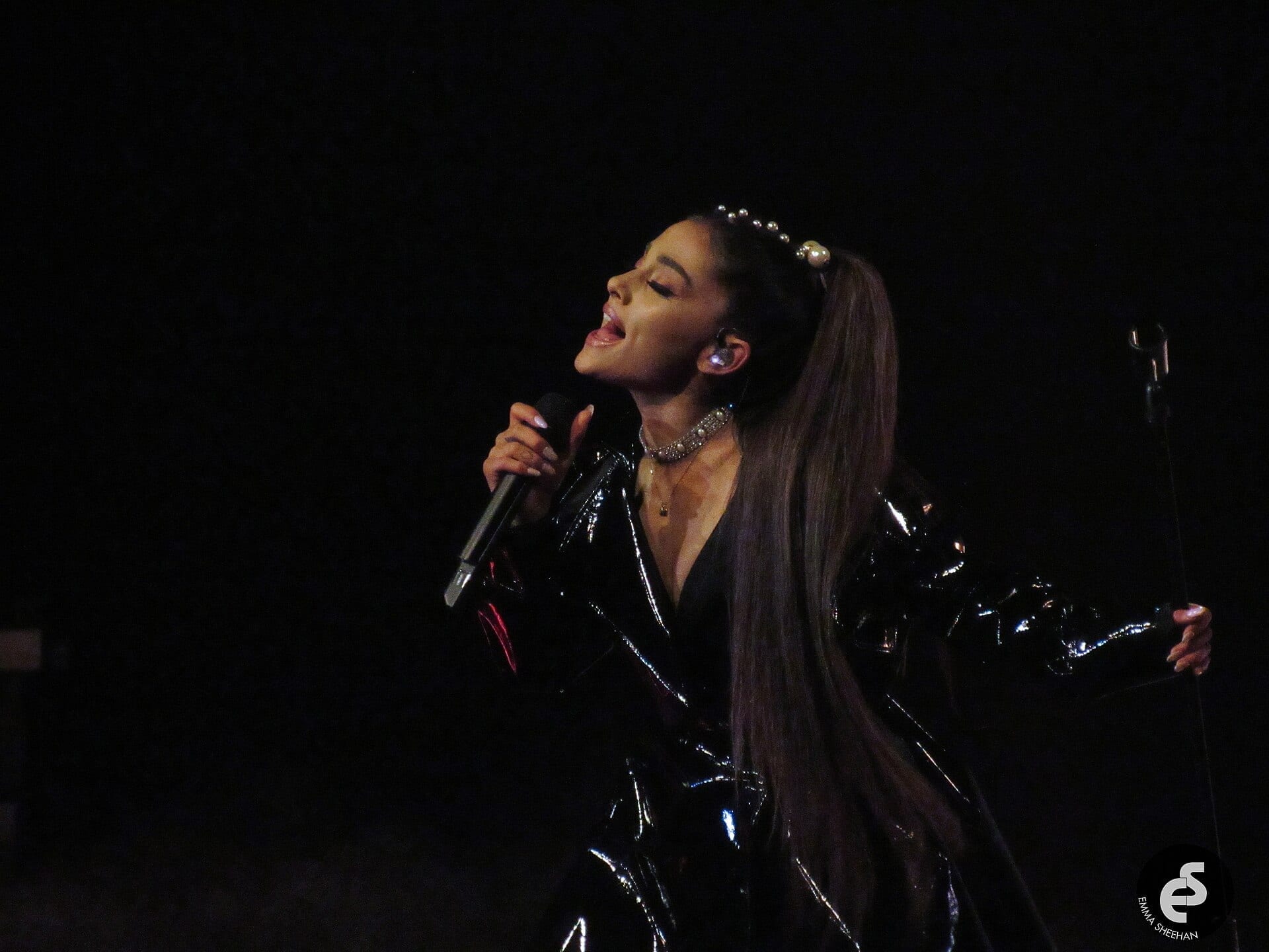 Ariana Grande sings into a handheld microphone on a dark concert stage, head tilted back, wearing a black patent jacket and a pearl headband over a high ponytail.