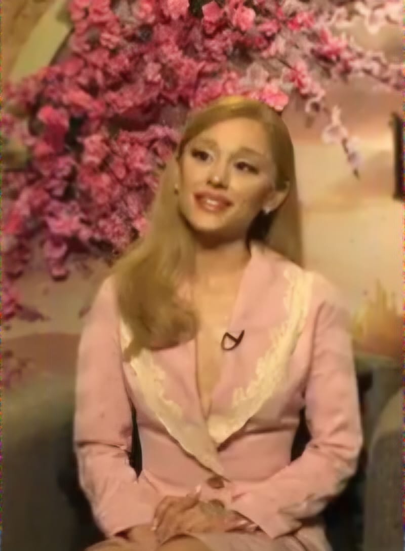 Ariana Grande, in a pink and cream jacket, smiles during an interview in front of a pink floral backdrop.