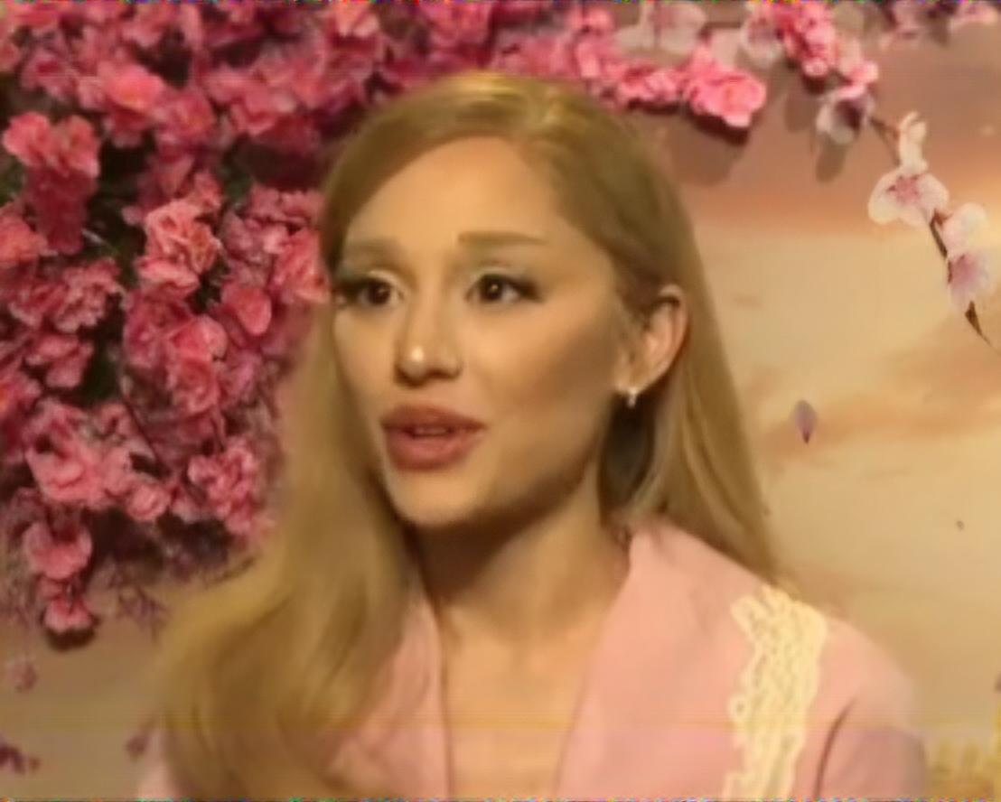 Ariana Grande talks mid-interview in a pale pink jacket, blonde hair down, against a backdrop of pink blossom.