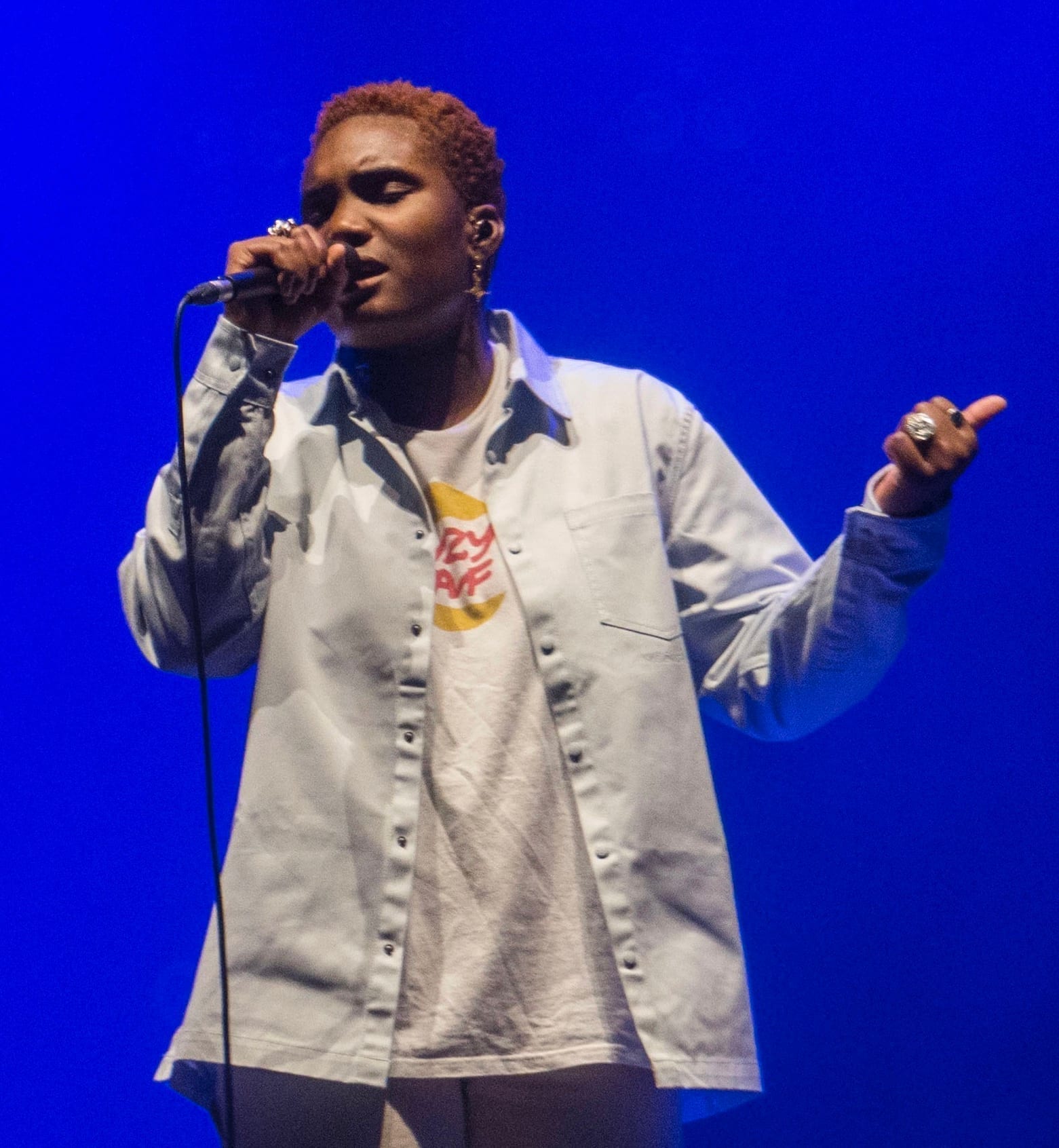 Arlo Parks sings into a microphone on stage lit in deep blue, wearing an open white shirt over a graphic t-shirt and pointing with her other hand.