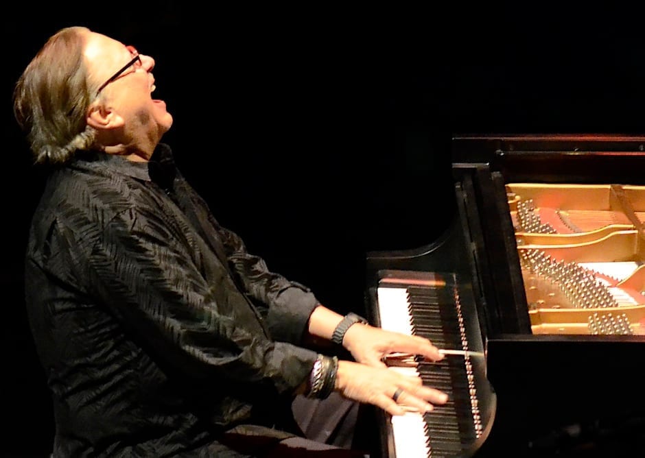 Arturo Sandoval, head tilted back, plays a grand piano on a dark stage, wearing a black patterned shirt and layered bracelets.