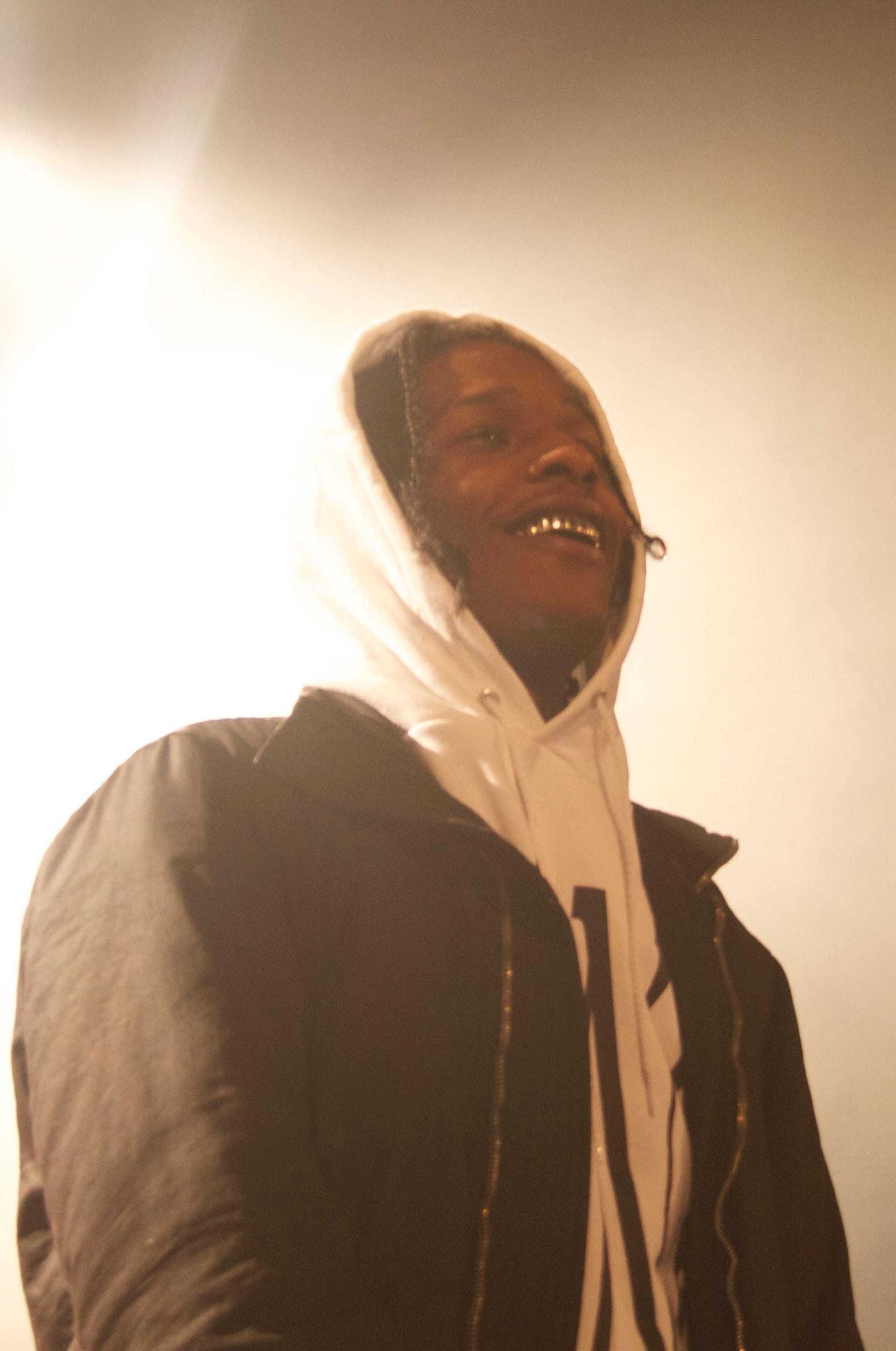 A$AP Rocky smiles widely, showing a gold grill, while wearing a white hoodie under a dark jacket, backlit by bright stage light.